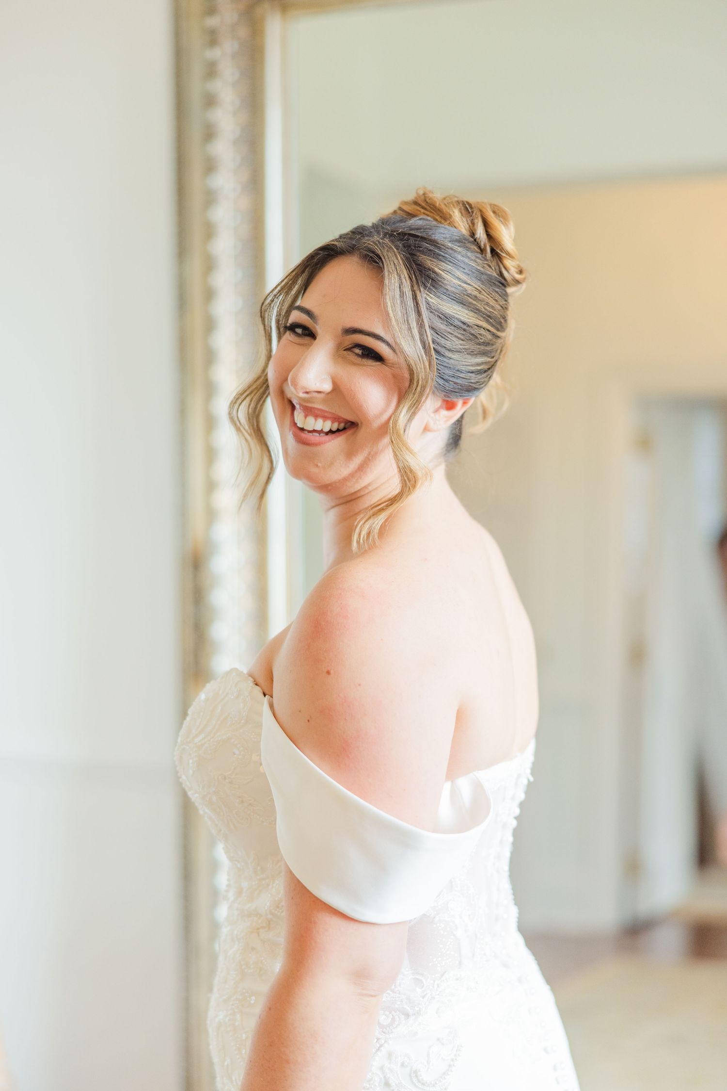 A bride in a white wedding dress smiles joyfully while getting ready in a bright sunlit room.