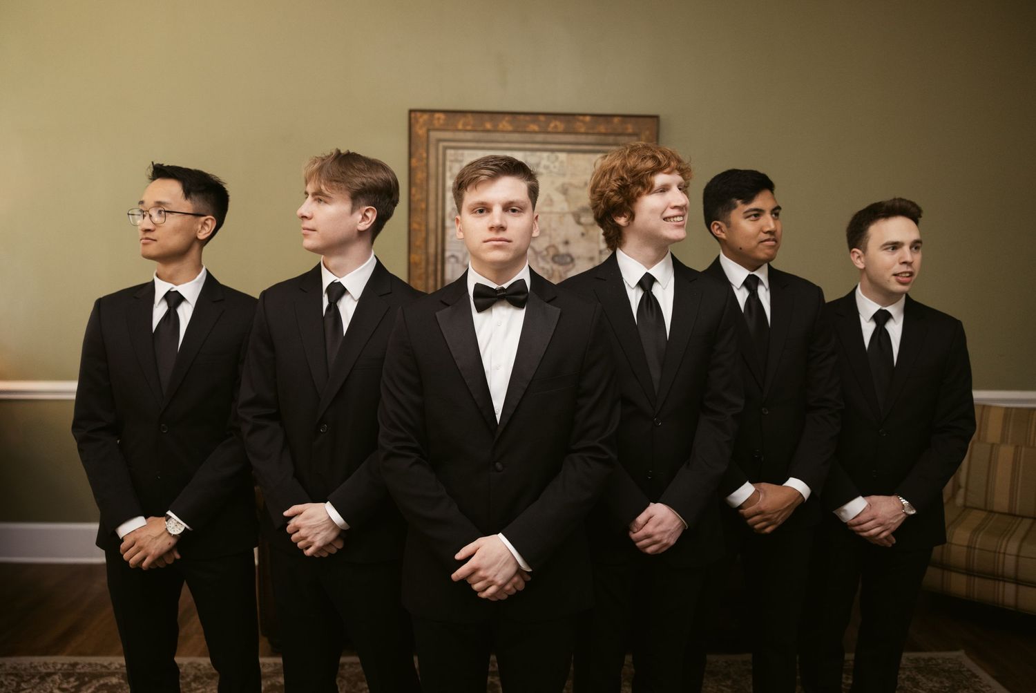Groomsmen in black tuxedos lined up against a dark wall for formal wedding photos.
