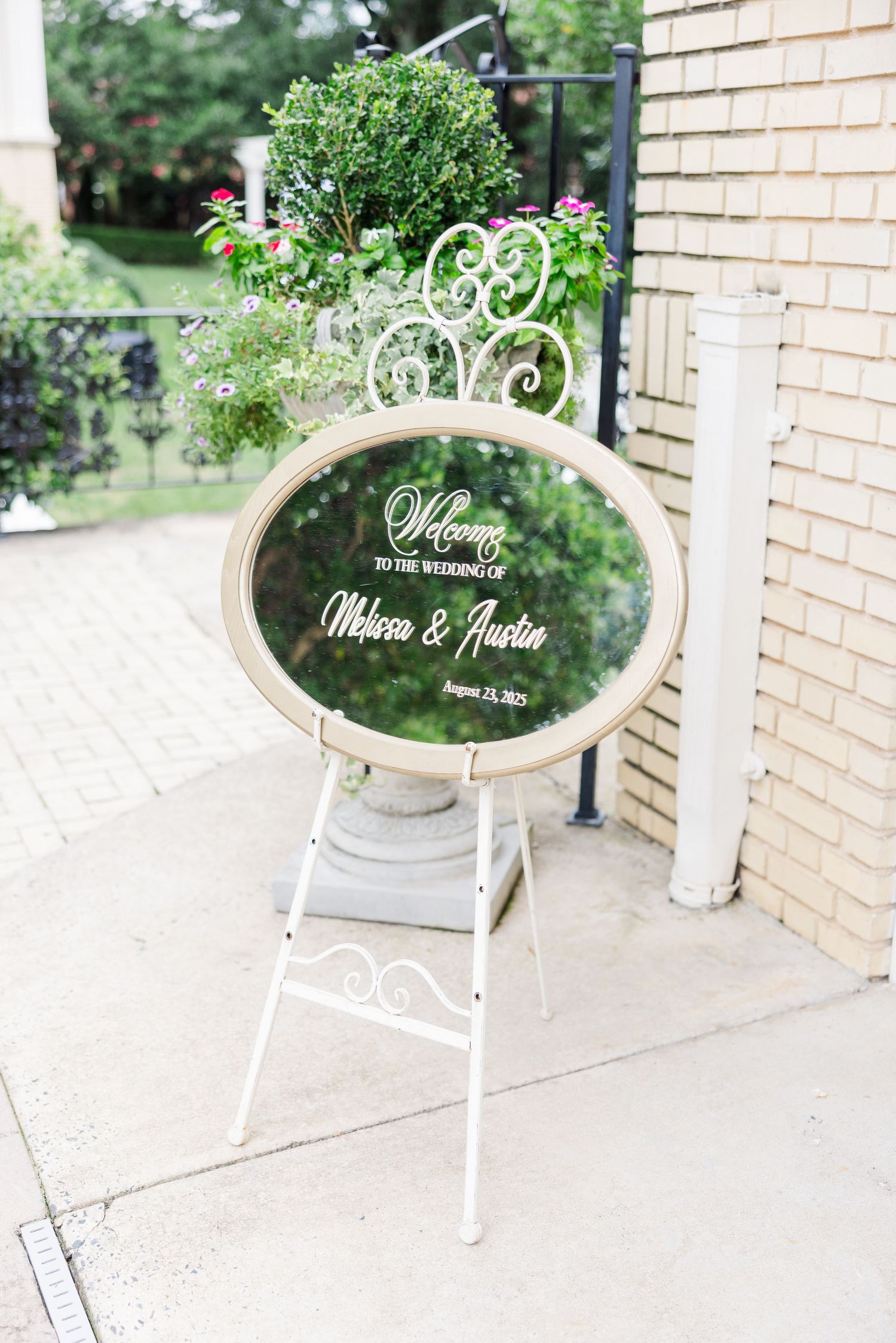 Elegant white decorative wedding welcome sign with ornate frame displayed outdoors against a brick wall.