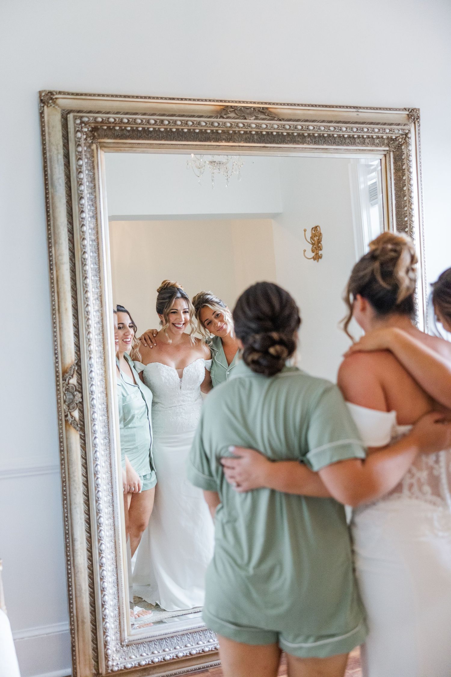 Bridesmaids in mint green dresses help with wedding preparations in front of an ornate mirror.