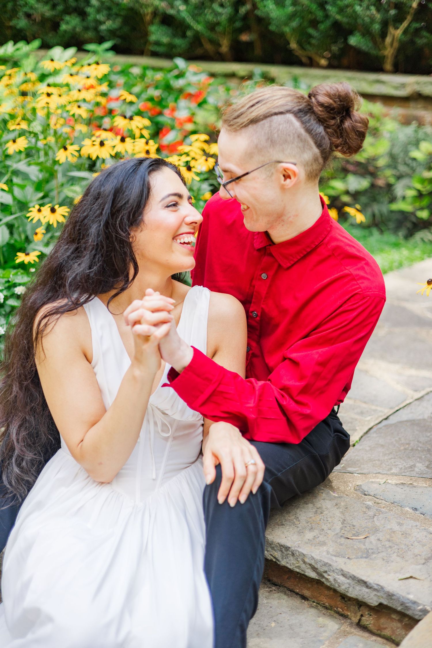 A joyful couple laughs together while sitting on stone steps surrounded by yellow flowers.
