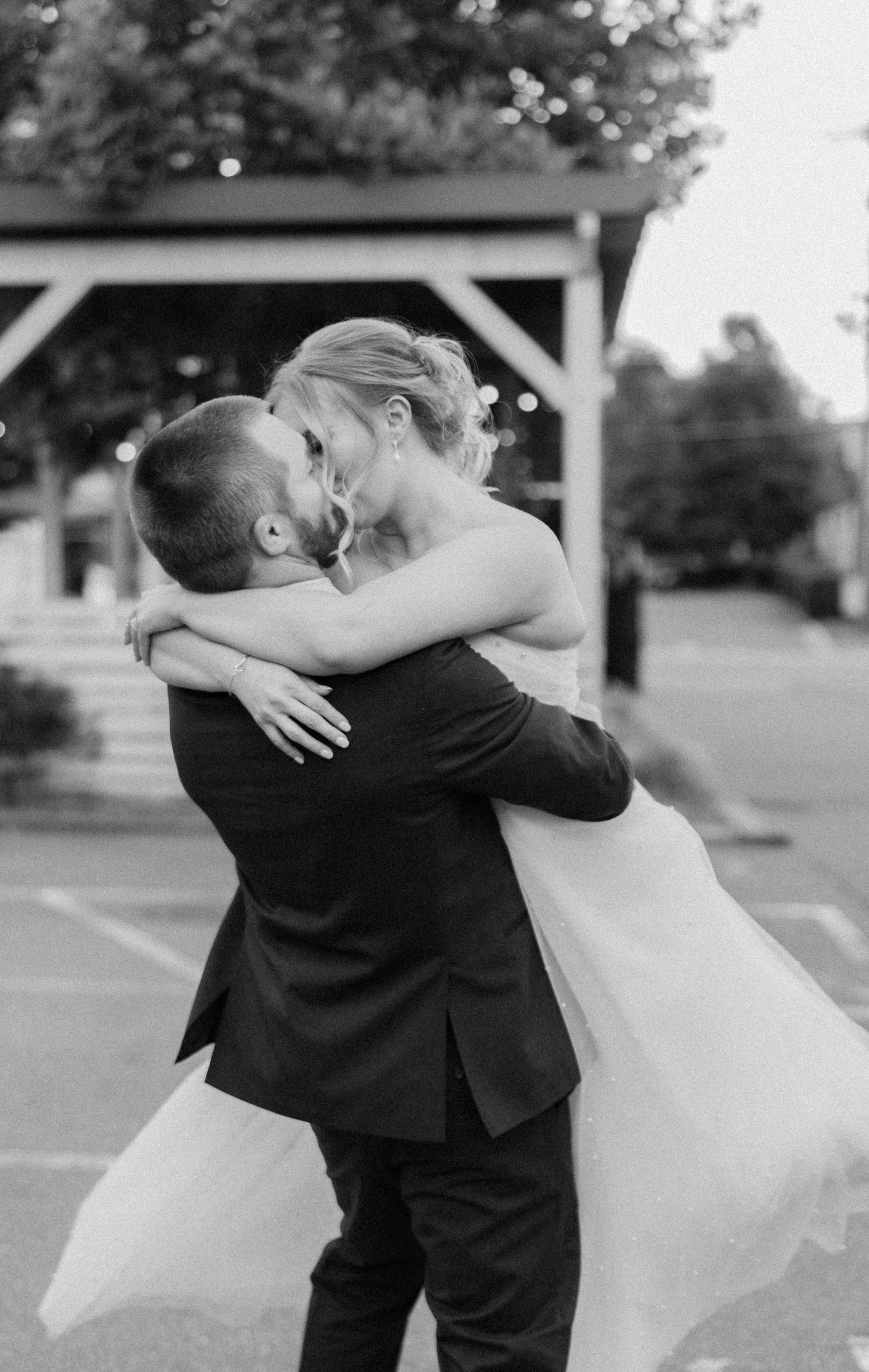 A black and white wedding photo captures an intimate embrace between newlyweds outdoors.