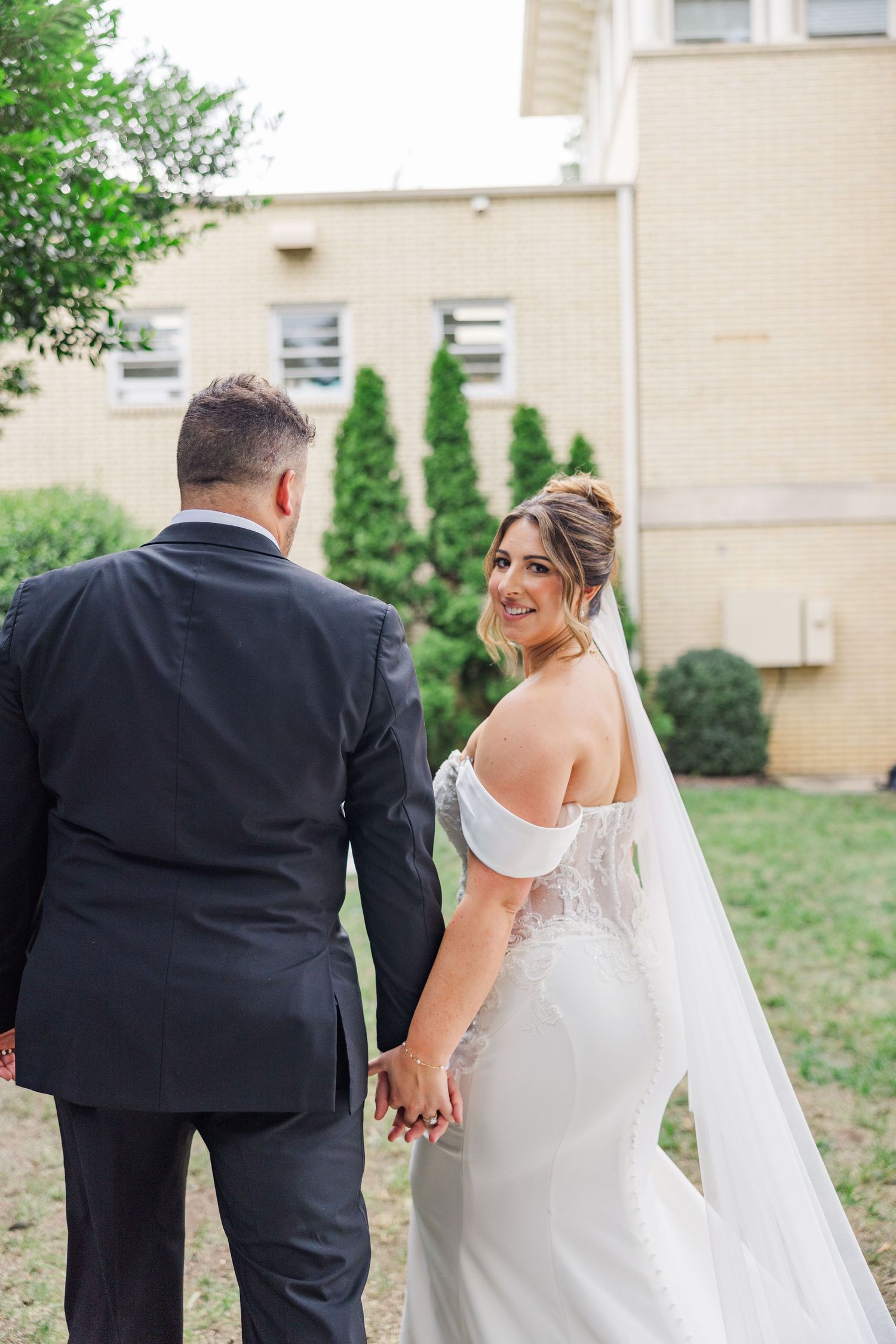 Newlyweds share intimate moment while walking outdoors near beige building.