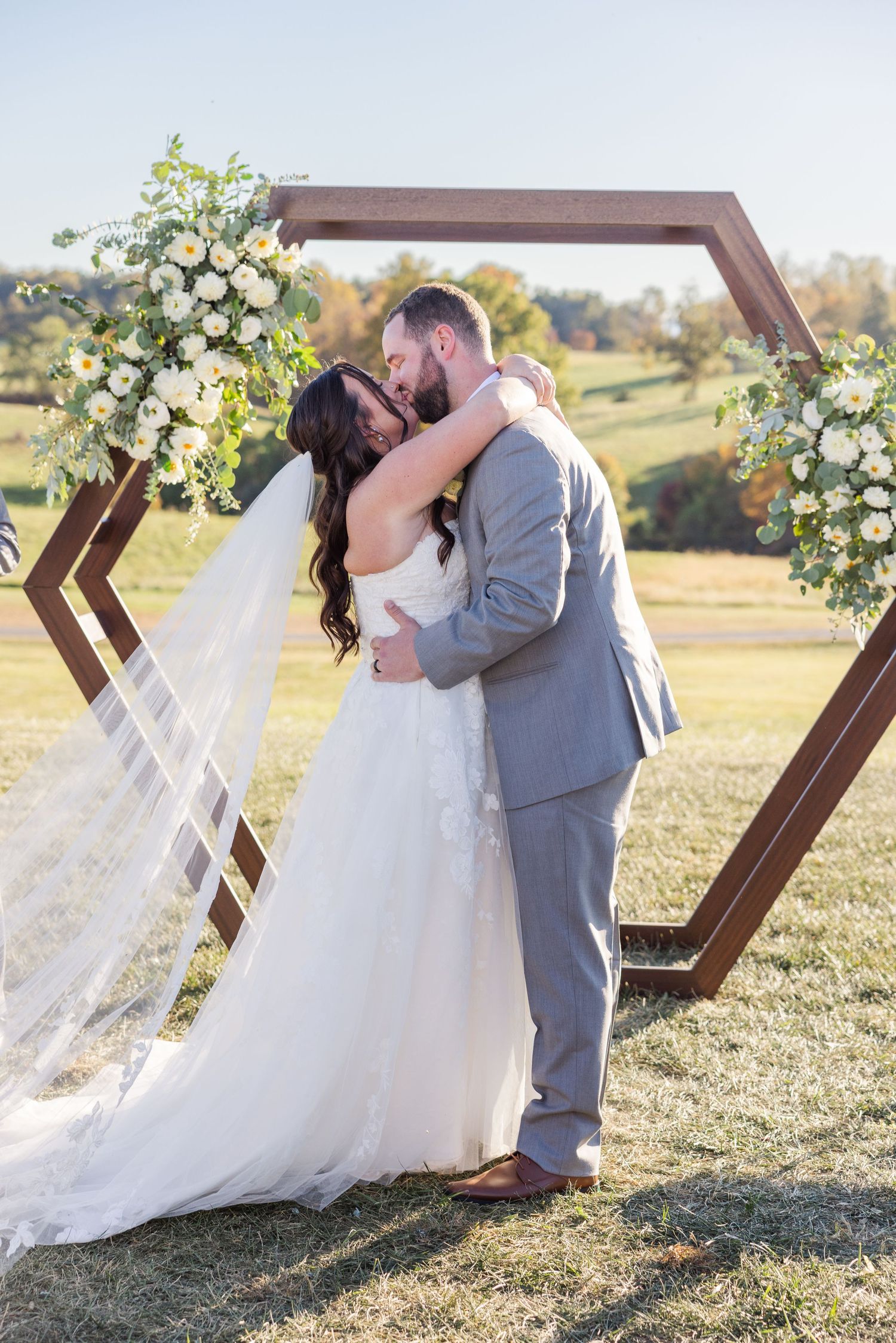 A romantic wedding couple shares a kiss under a wooden arch decorated with white flowers in an outdoor field setting.