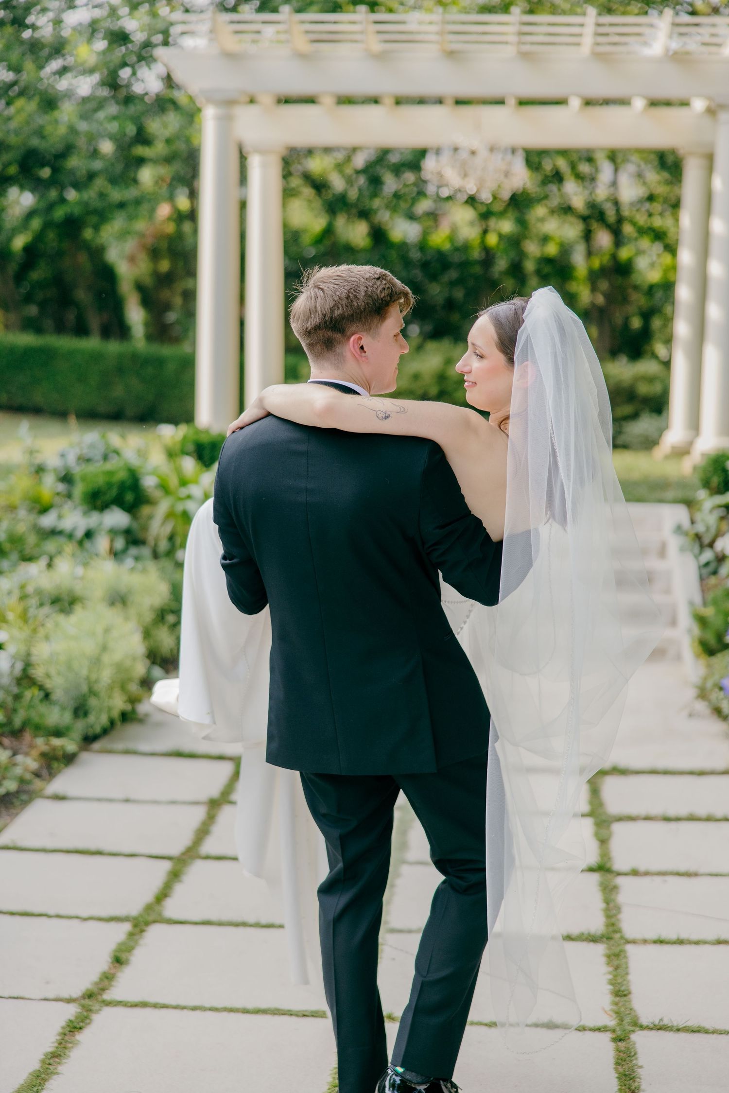 A wedding couple dances together on an outdoor stone patio surrounded by lush gardens.