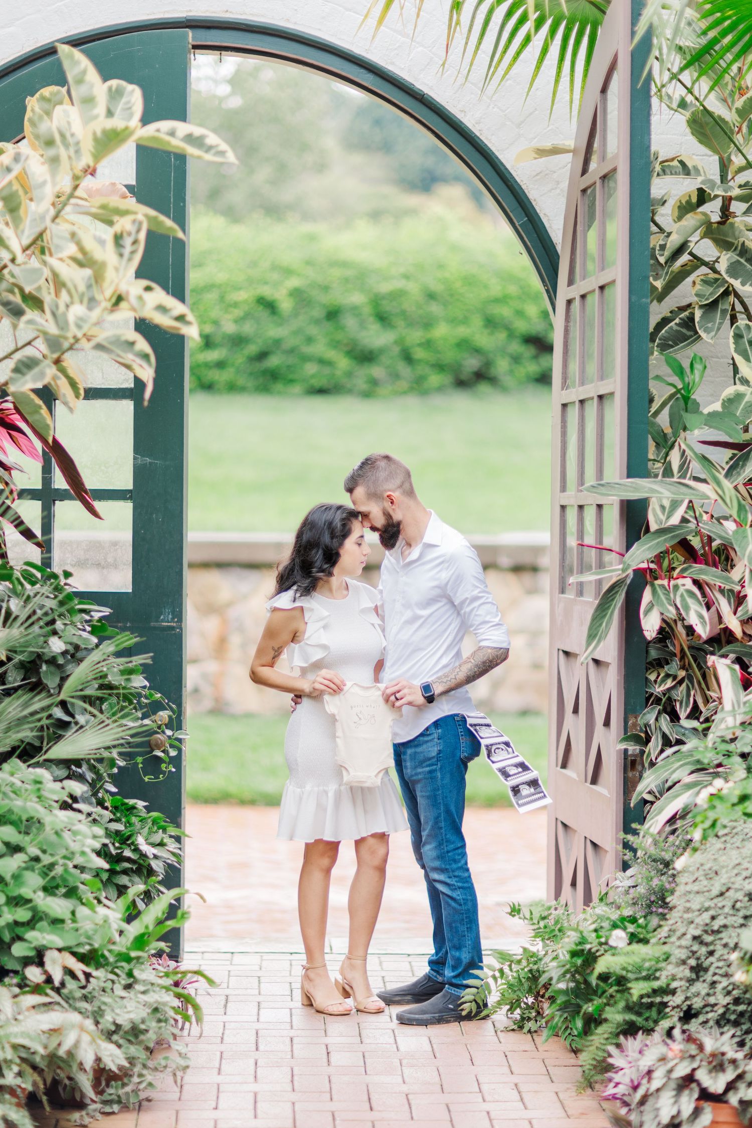 A romantic couple poses beneath a garden archway surrounded by climbing plants and flowers.