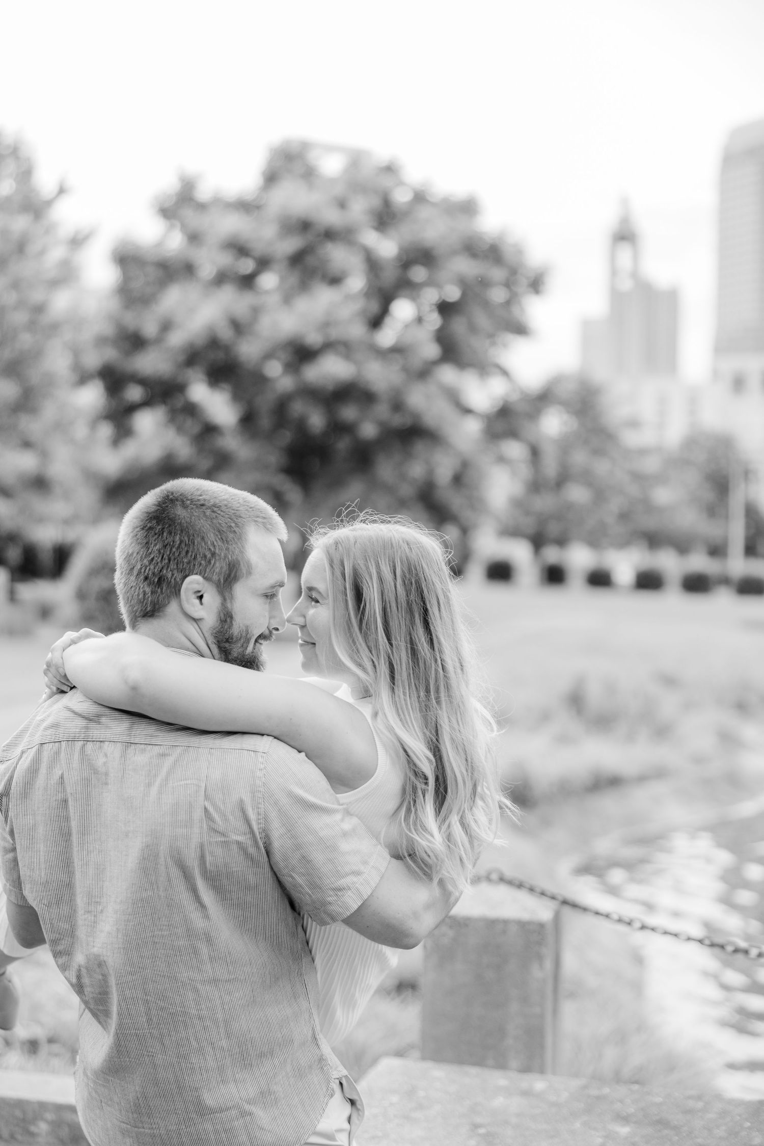 A black and white portrait of a couple embracing near water with city architecture visible.