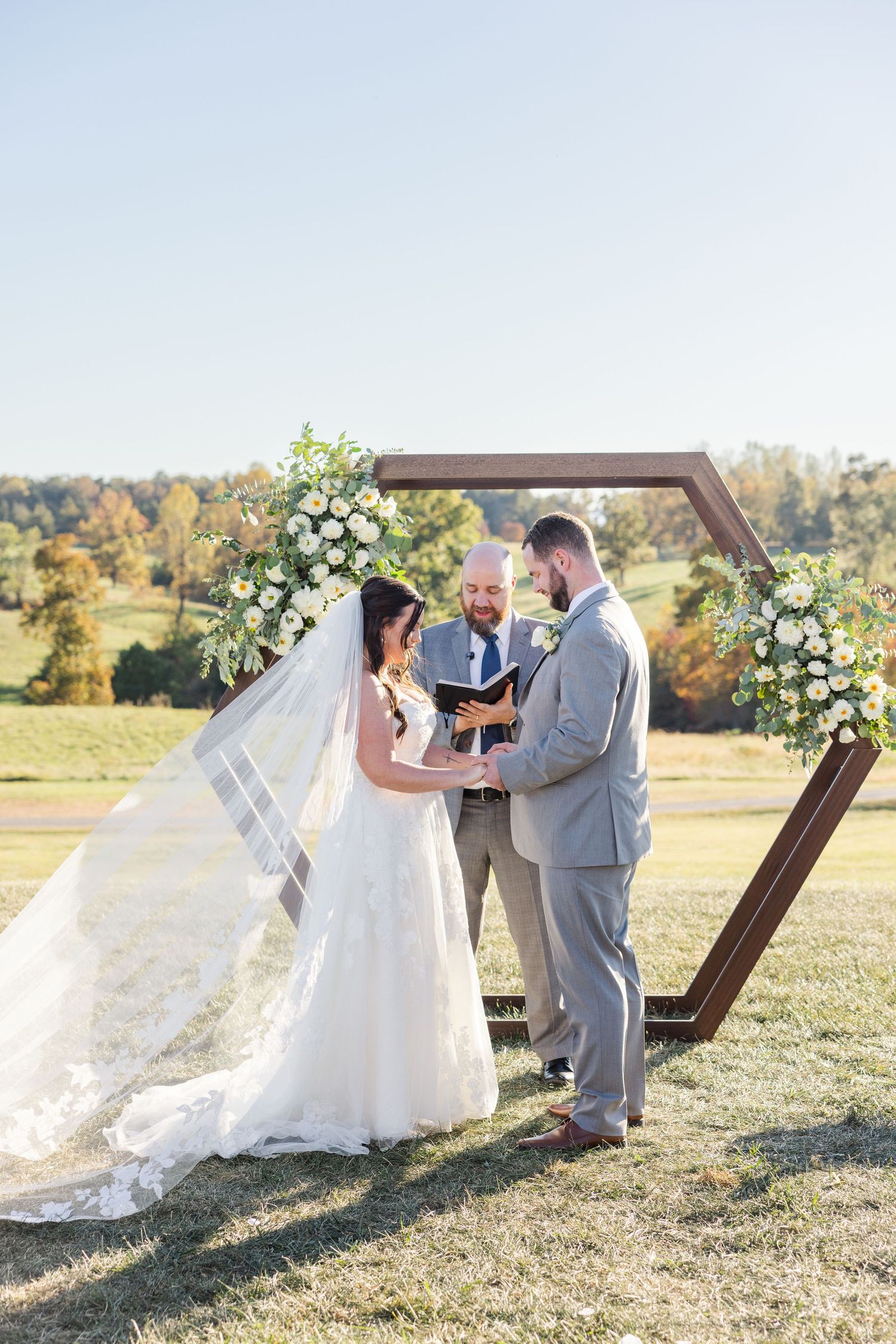 A romantic outdoor wedding ceremony takes place under a wooden hexagonal arch decorated with white flowers.
