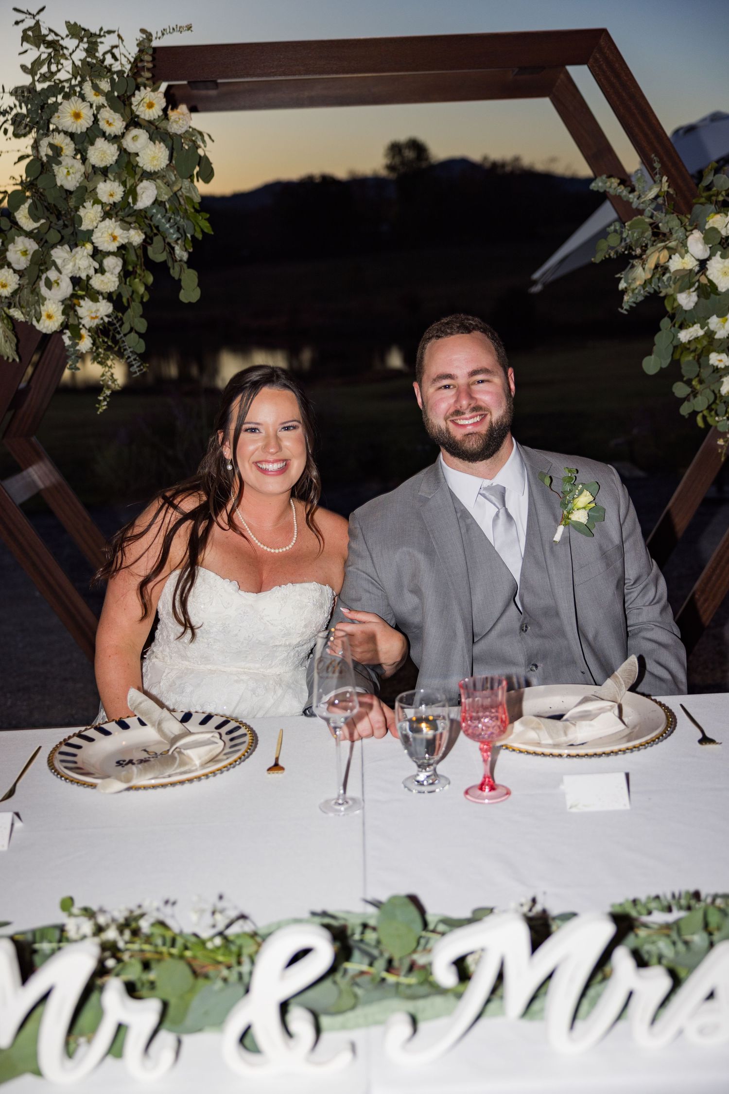 A couple sits at an elegant wedding reception table decorated with white floral arrangements and glassware at sunset.