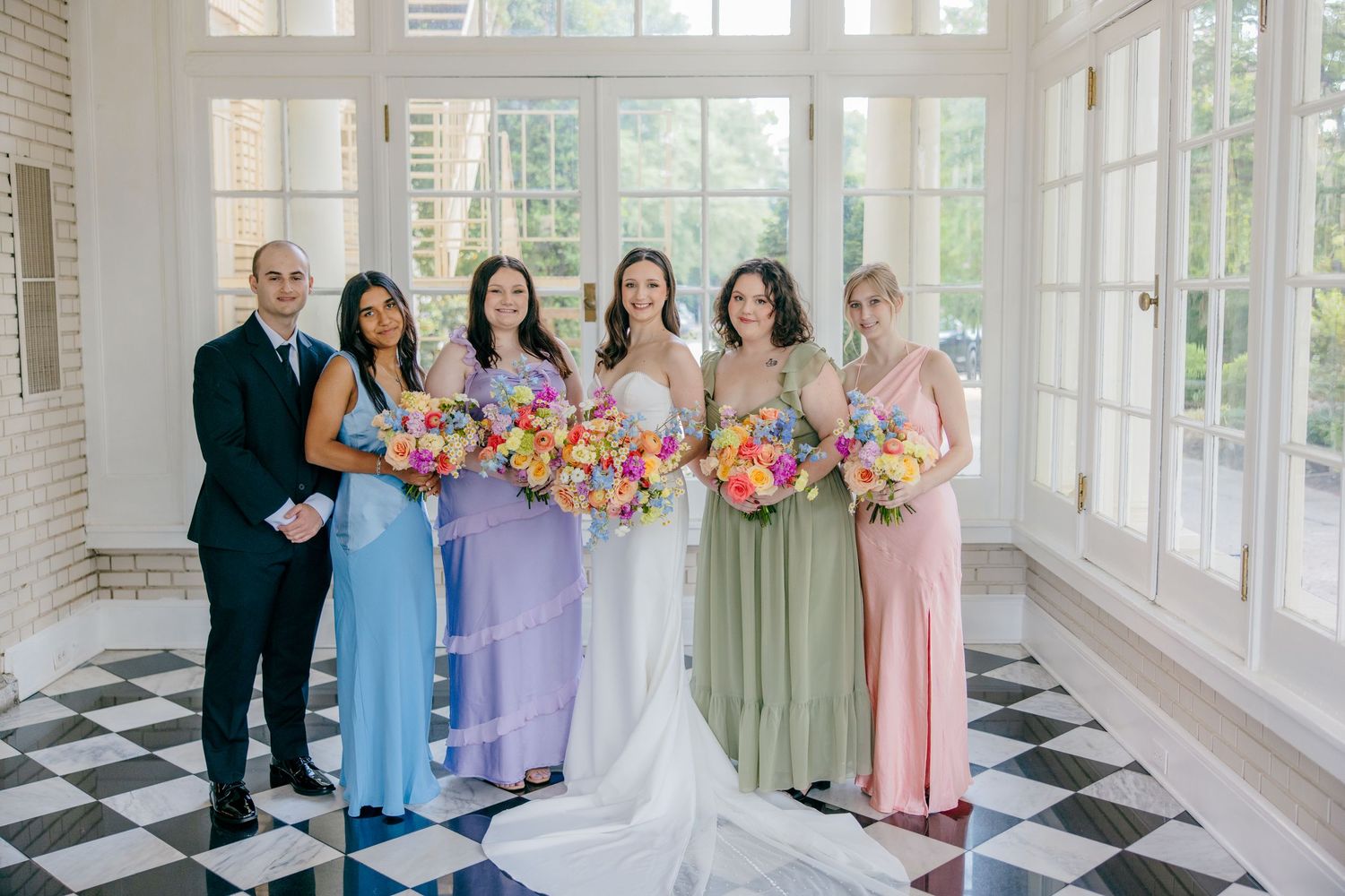 Wedding party in pastel dresses posing in a bright sunlit room with checkerboard floors.