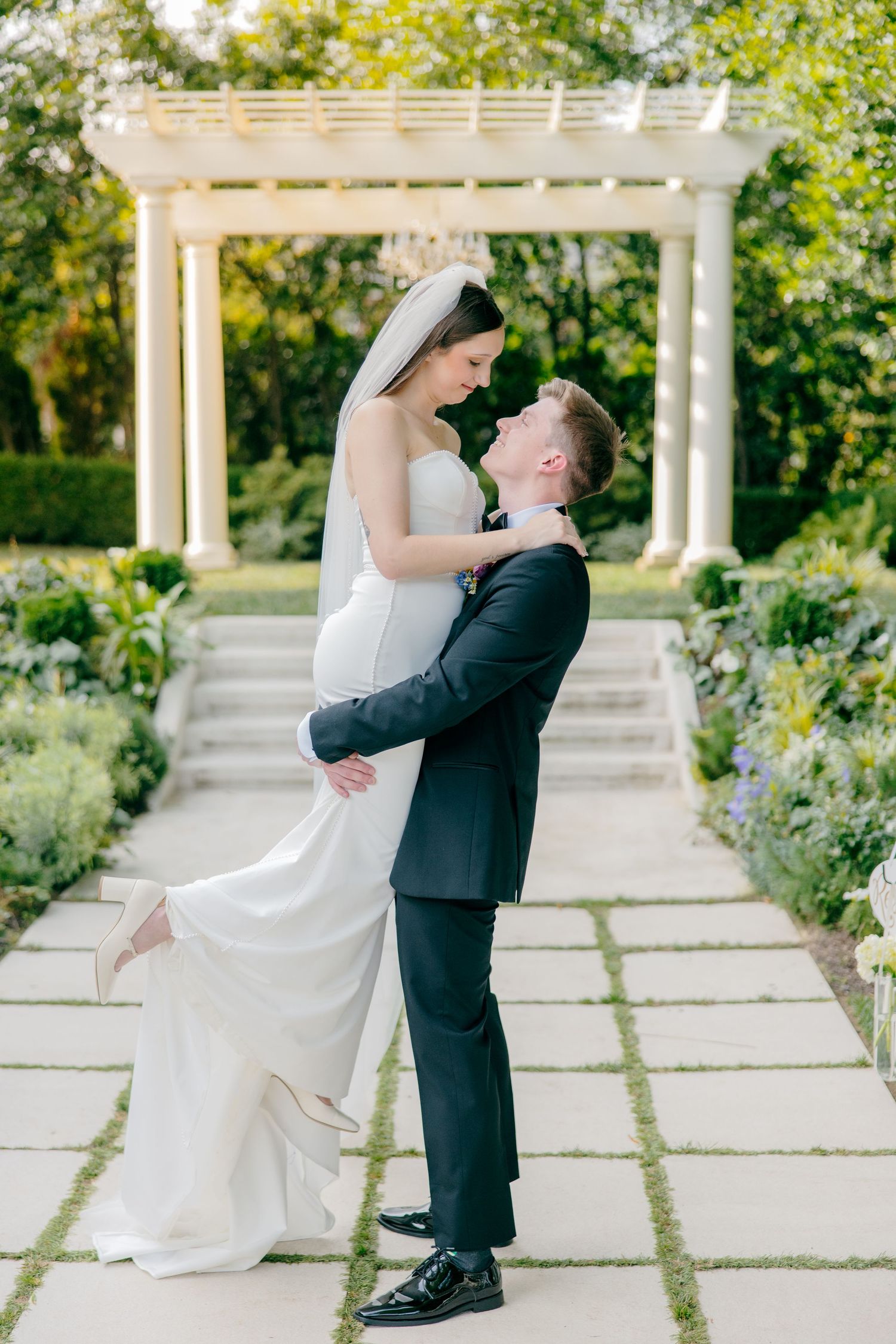 Romantic wedding portrait under a white pergola surrounded by lush garden landscaping.