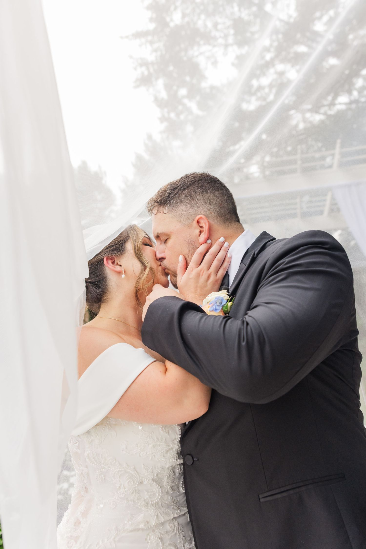 Romantic wedding kiss moment captured through sheer white curtains.