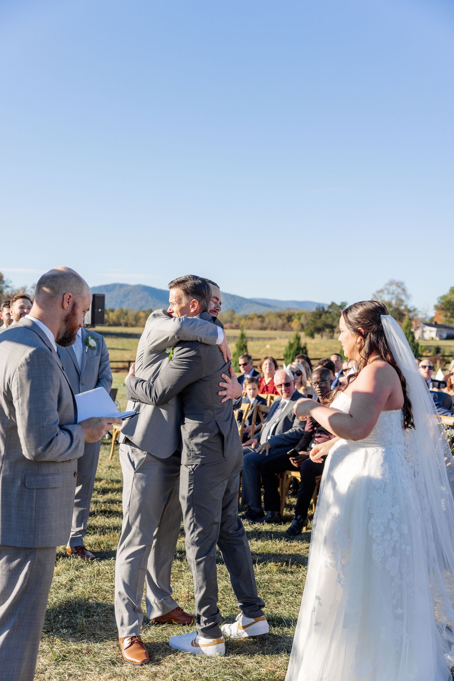 Wedding party in gray suits and white dresses gathered outdoors under blue sky.