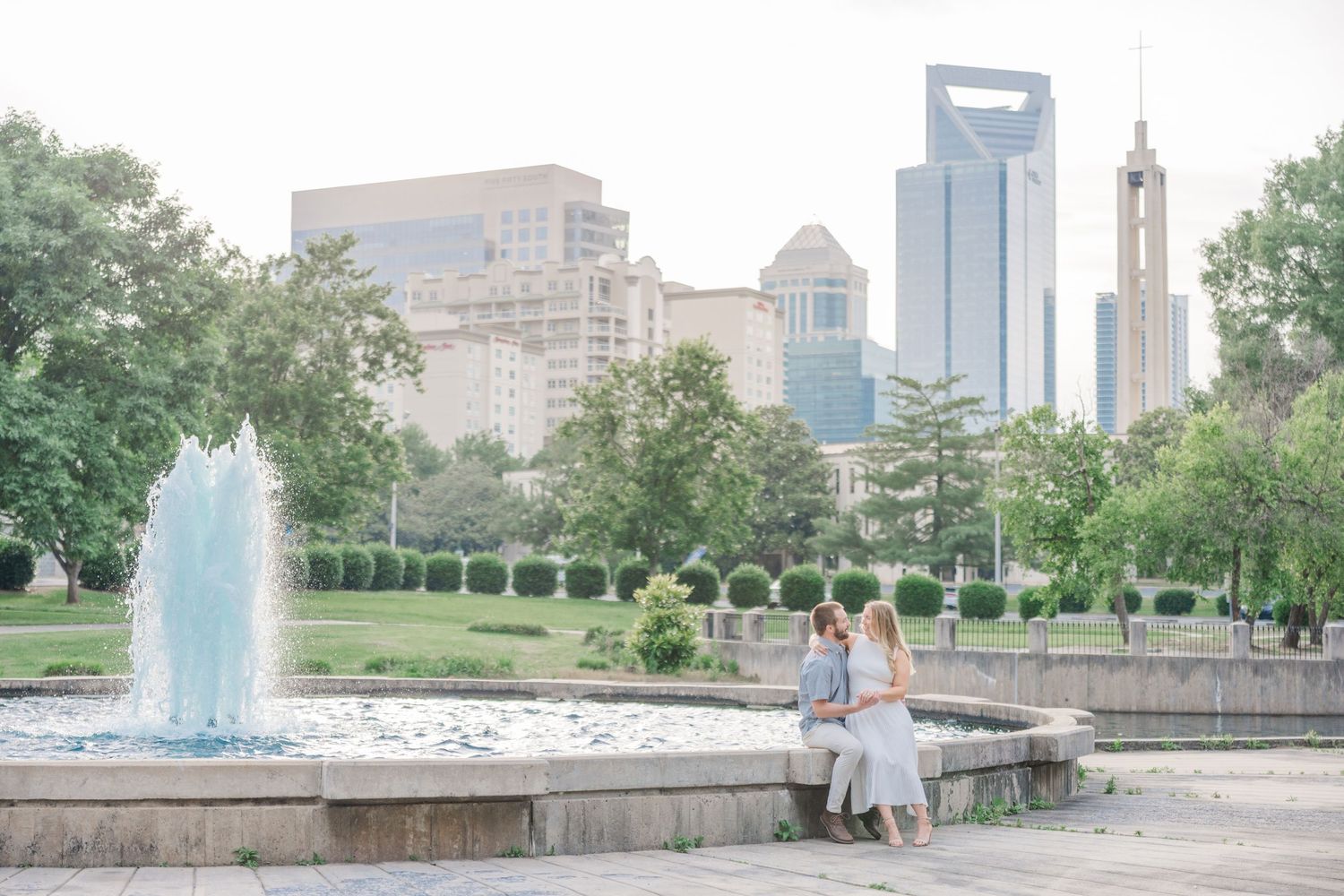 A couple poses for engagement photos along a scenic urban park with fountains and skyline views in the background.