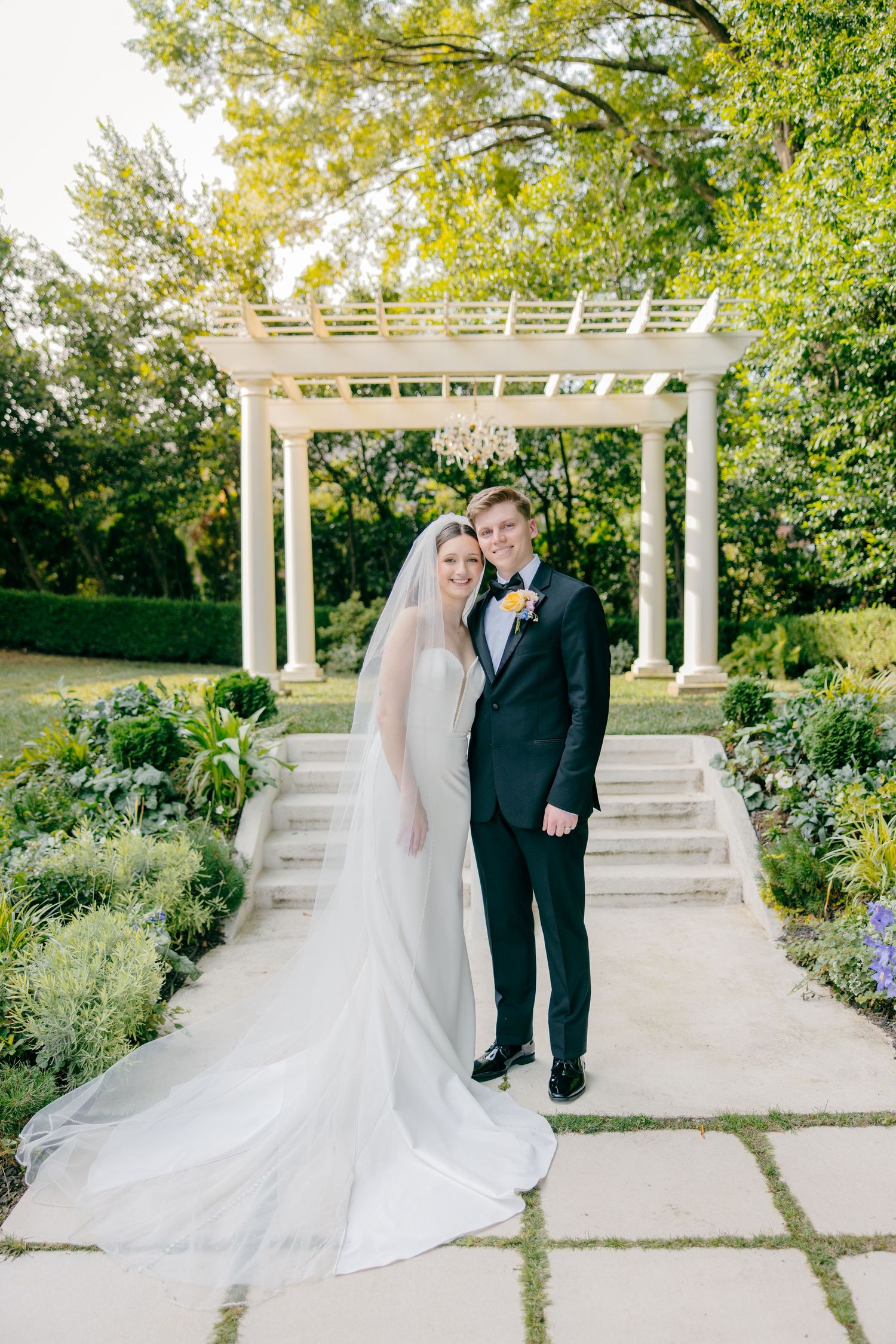 A wedding couple poses under a white pergola surrounded by lush garden landscaping.