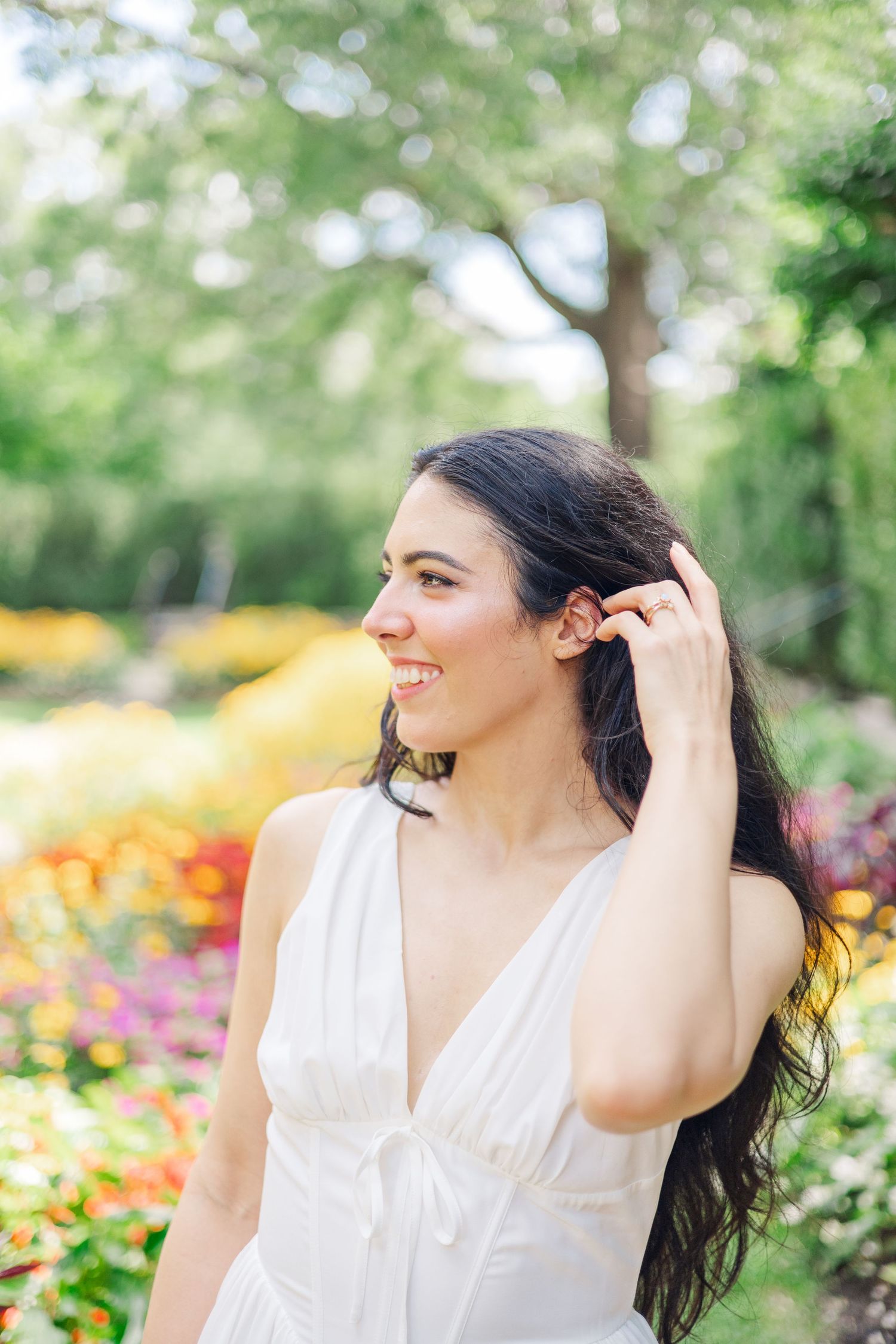 A person in a white dress smiles while posing in a sunny garden filled with colorful flowers.
