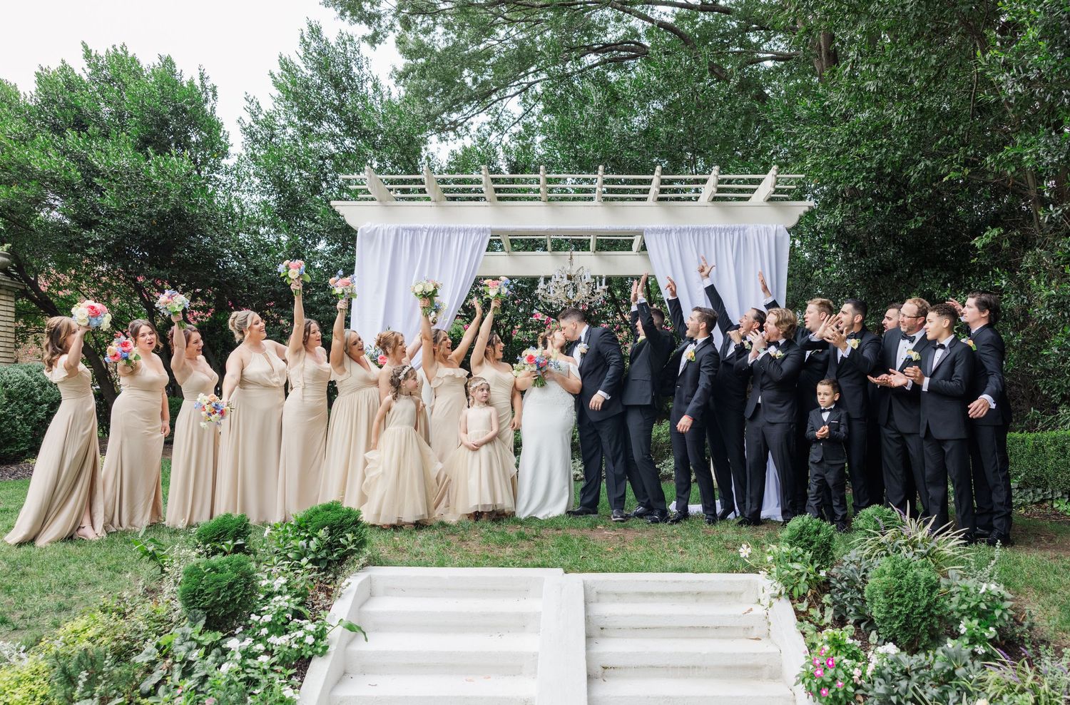 A wedding party celebrates outdoors with raised arms and joyful expressions in a garden setting.