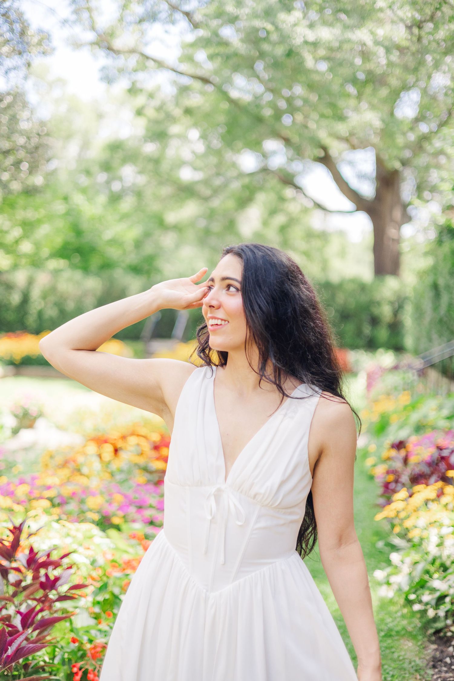 A woman in a flowing white dress poses gracefully in a vibrant garden filled with colorful flowers.