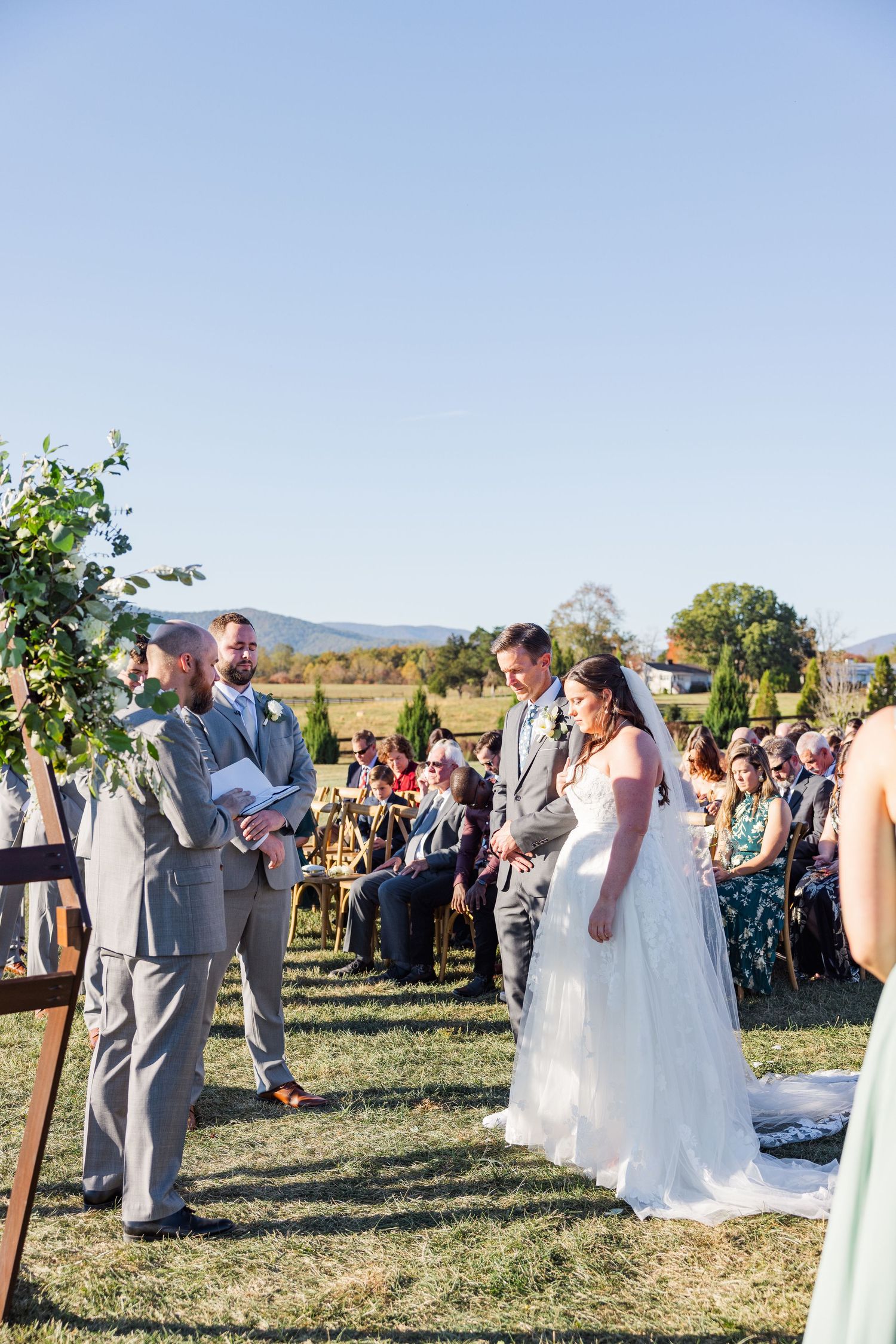 Outdoor wedding ceremony with guests gathered on a grassy field against a mountain backdrop.