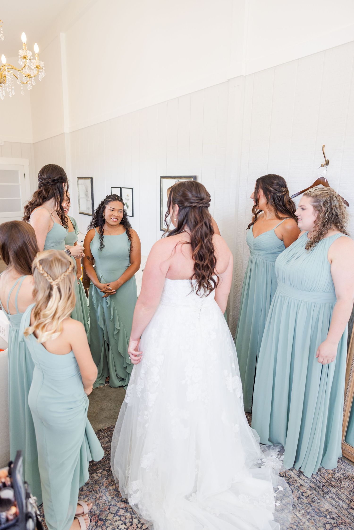 Bridesmaids in mint green dresses help the bride prepare in an elegant white room with chandelier.