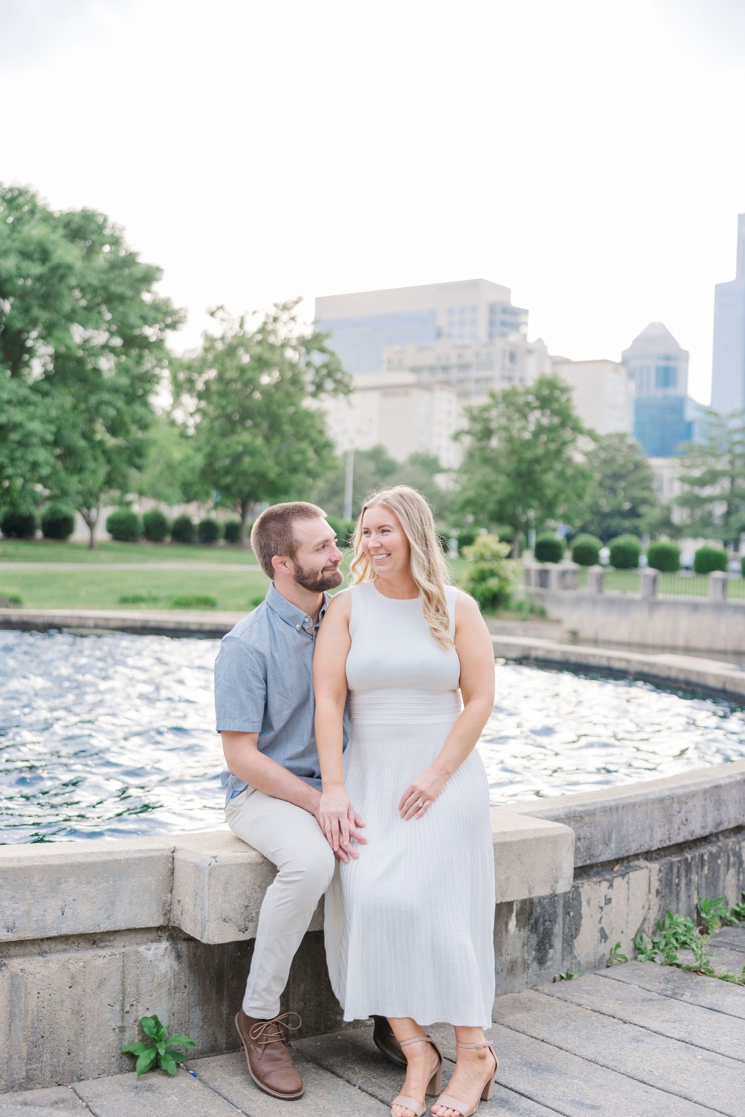A couple shares a romantic moment sitting on stone steps near a waterfront with a city skyline in the background.
