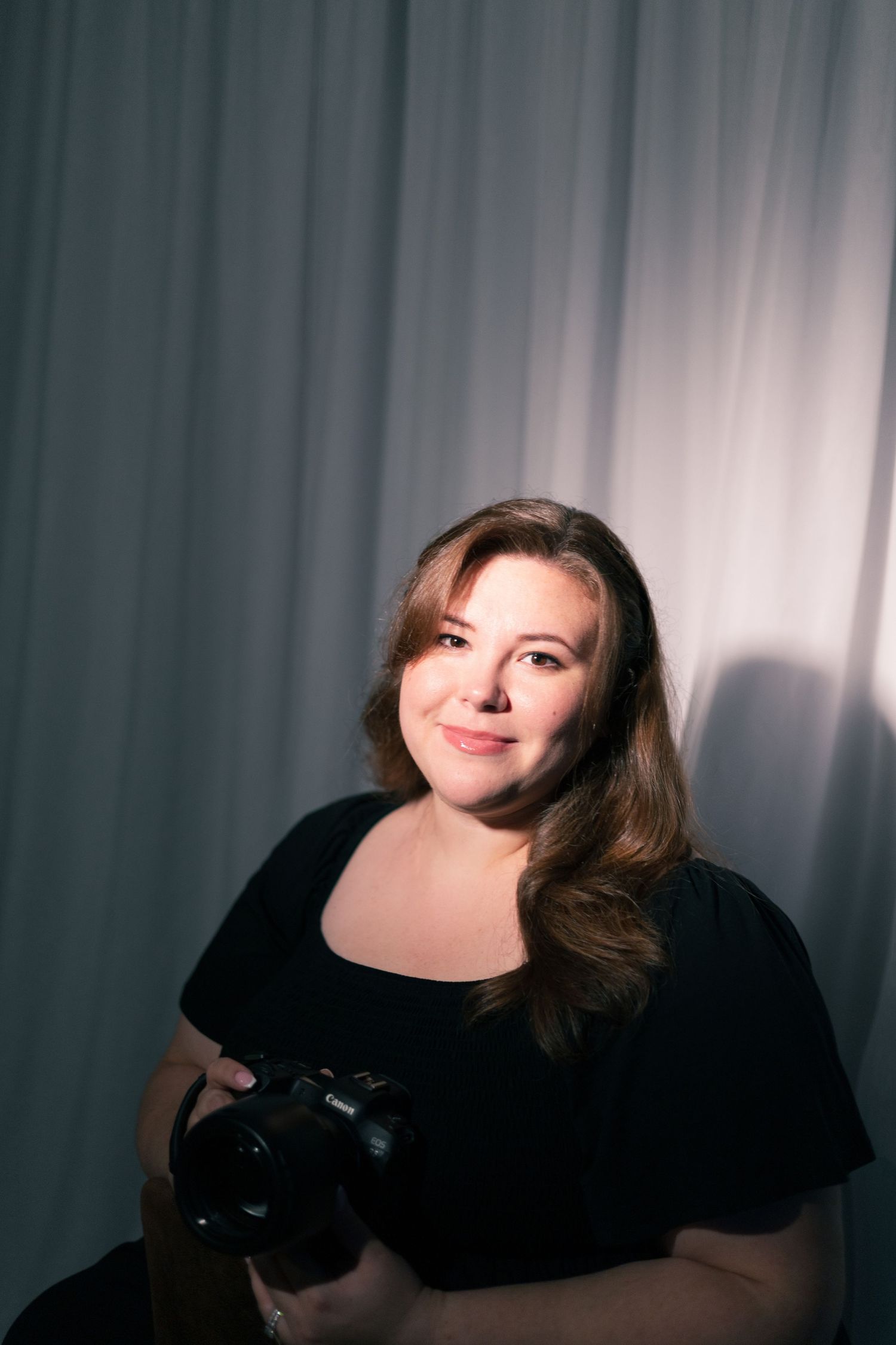 Woman in black top holding a camera, smiling against a gray curtain backdrop in a portrait photography setting.