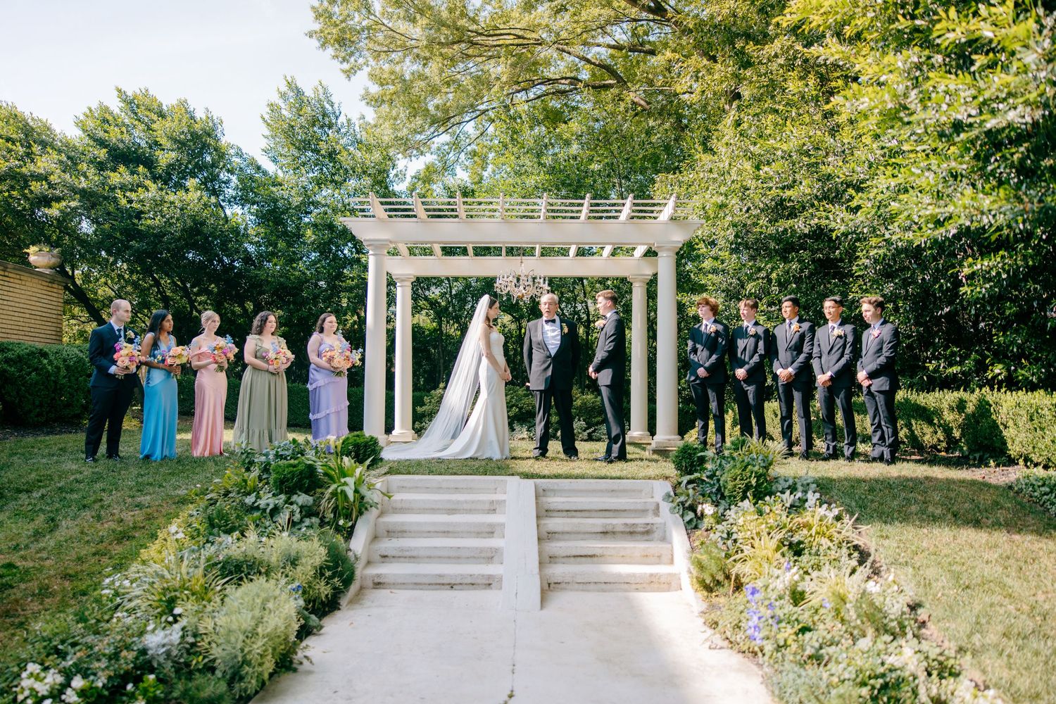 Wedding ceremony setup with white pergola and wedding party lined up in a garden setting.