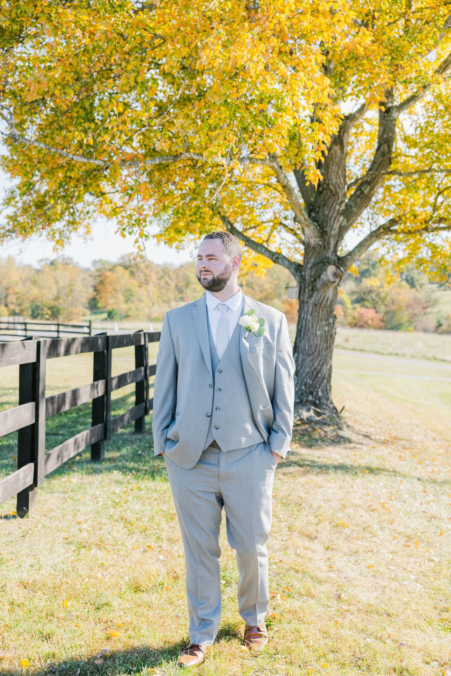 Groom in grey suit poses near a black fence with autumn trees displaying golden yellow leaves.