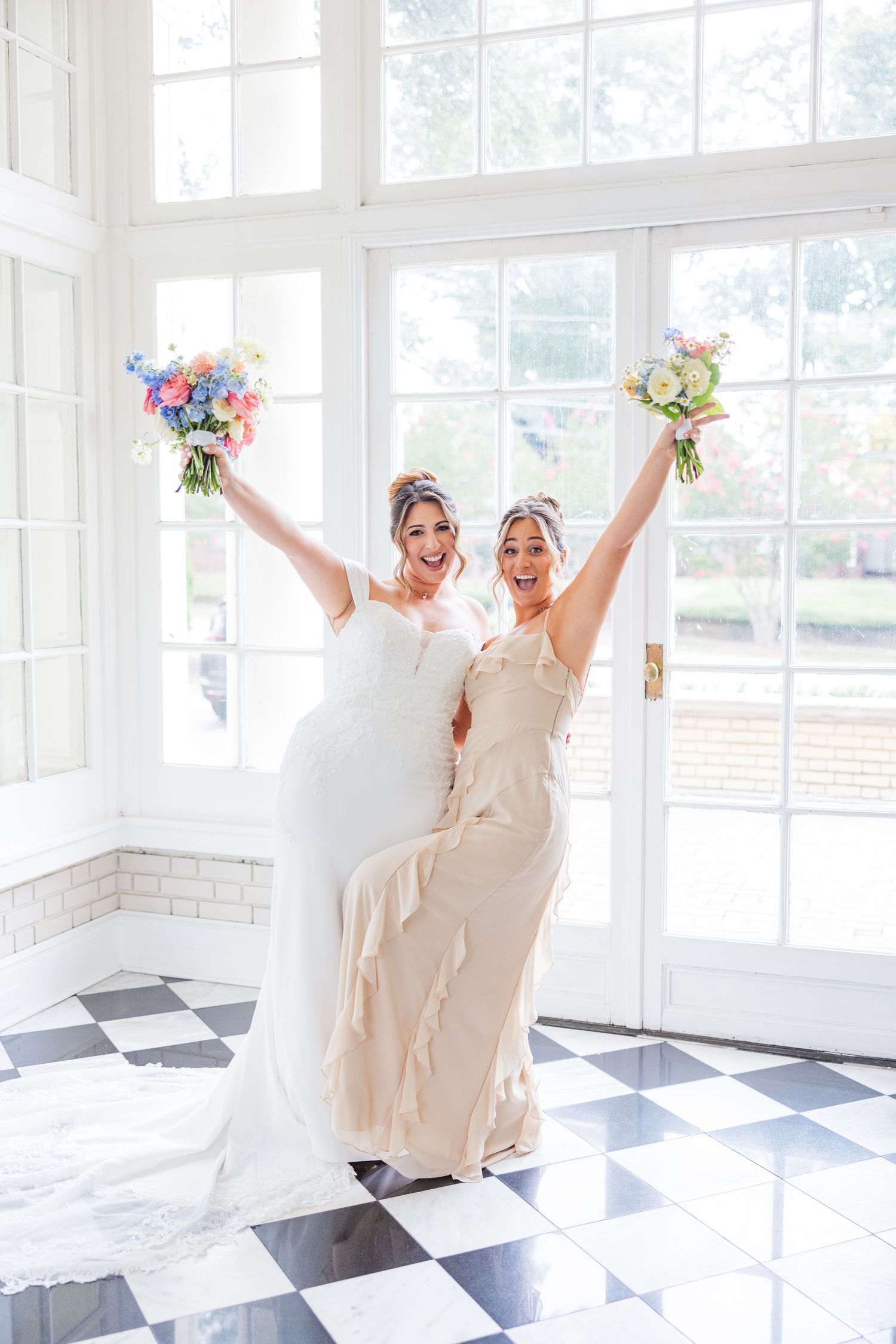 Two brides in white dresses celebrate joyfully on a black and white checkered floor with colorful bouquets.