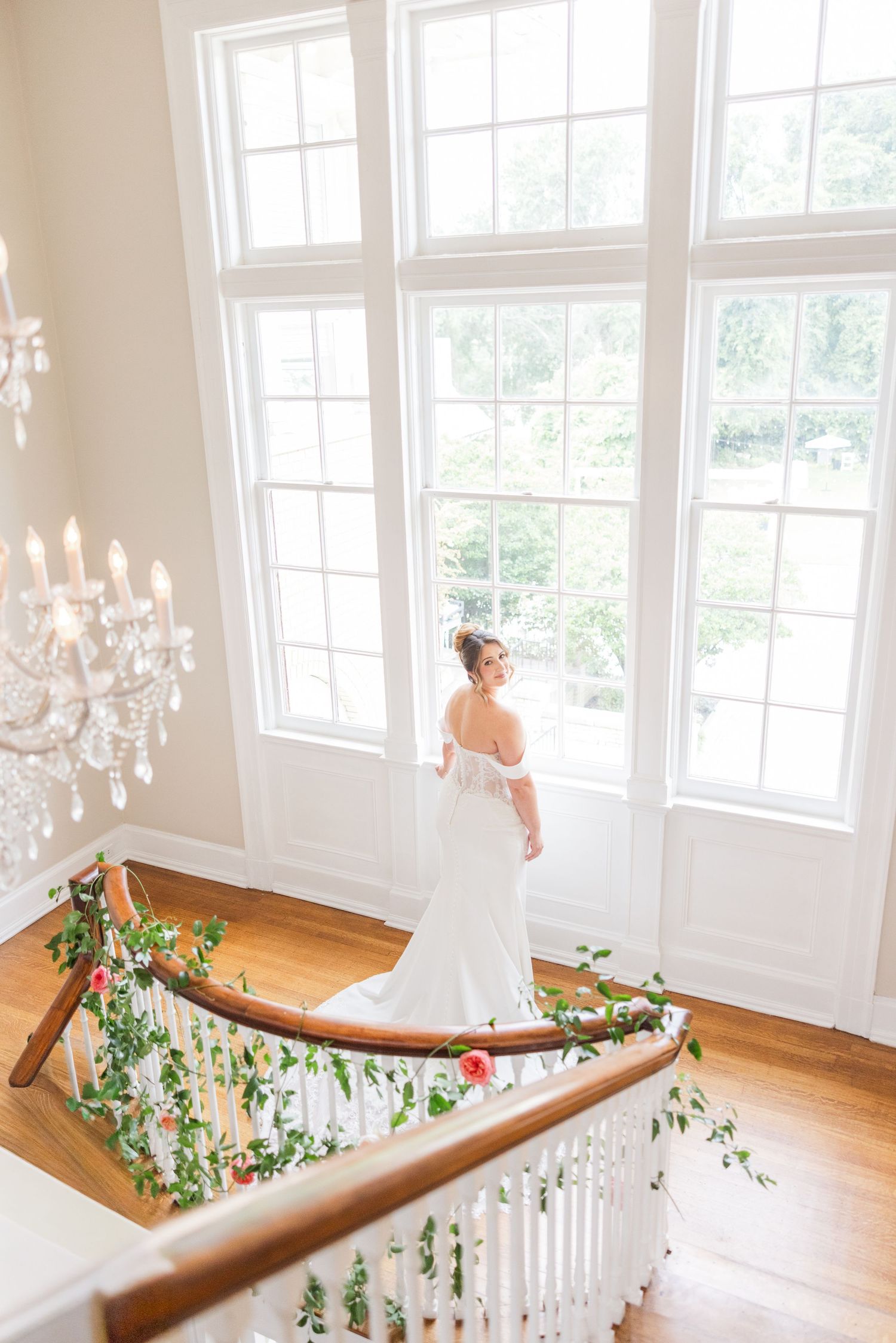 Elegant white staircase with crystal chandeliers and floral garland decorations in a bright, sunlit room.