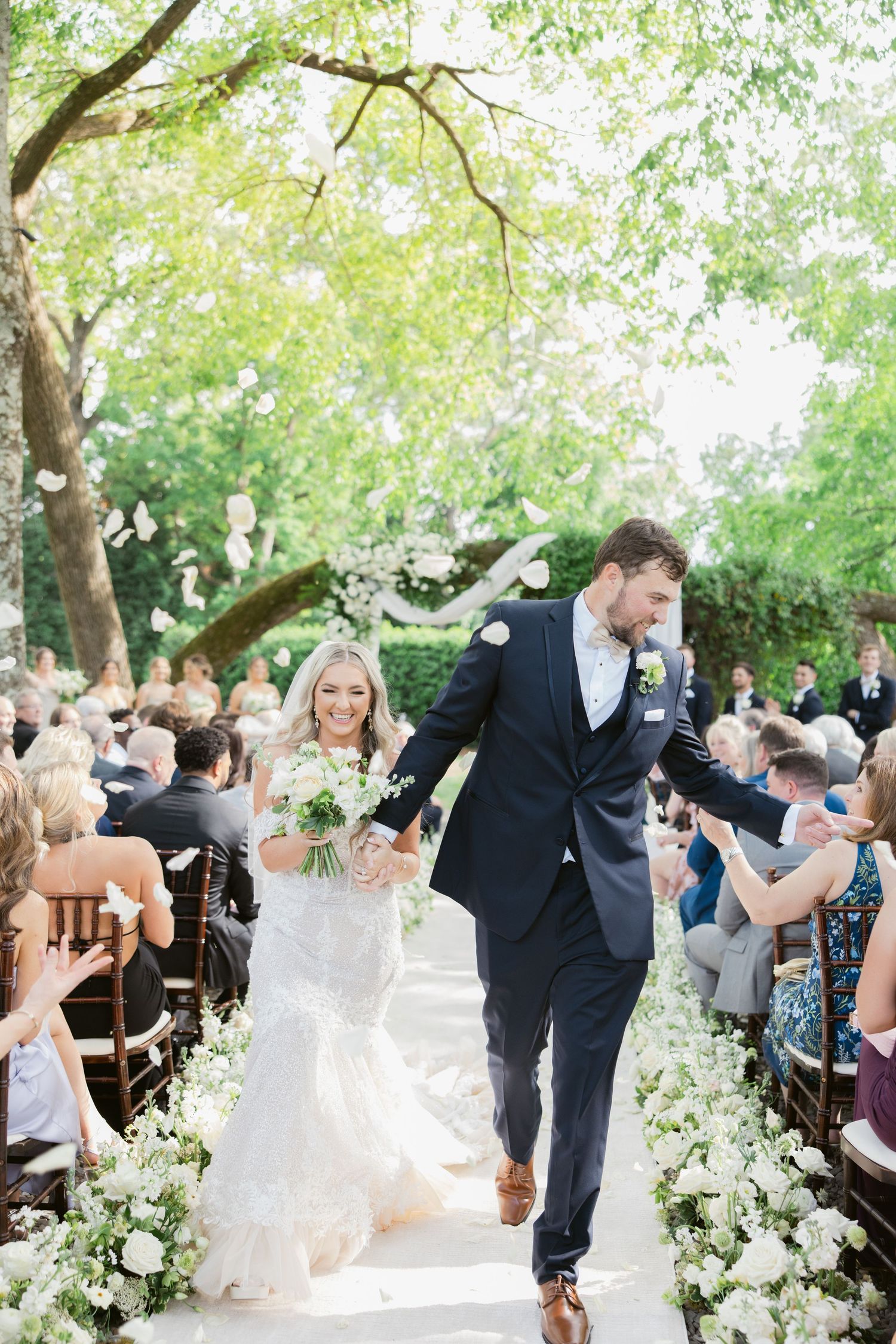 Newlyweds walk down the aisle surrounded by guests and white floral decorations in an outdoor garden ceremony.