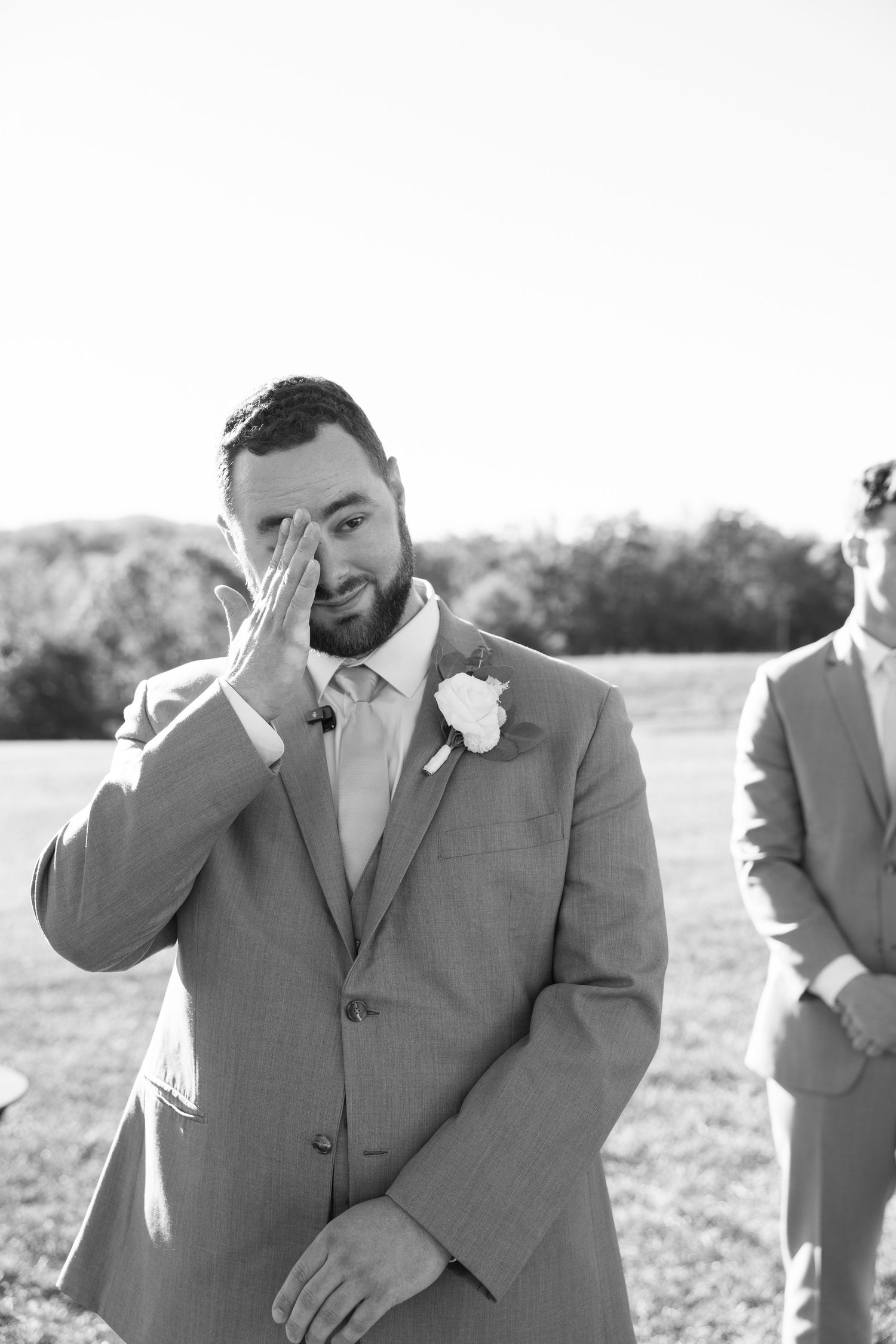 Groomsmen in gray suits share emotional reactions during outdoor wedding ceremony.