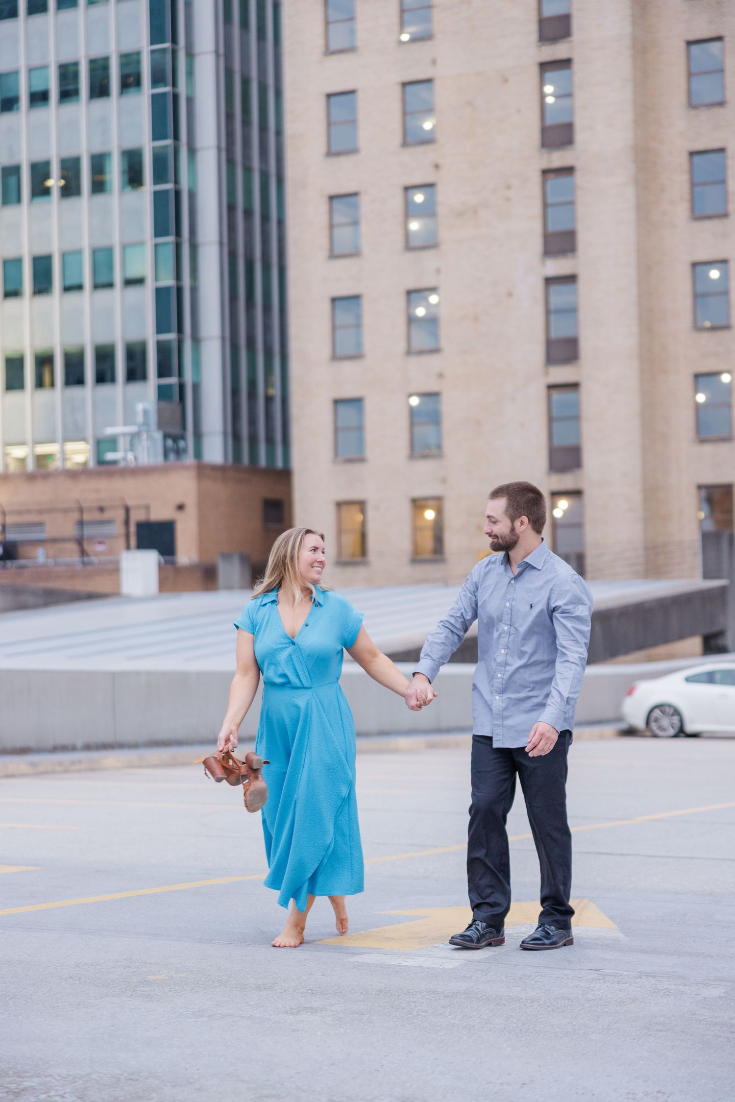 A couple in coordinated blue and gray outfits dance playfully in a parking lot with office buildings in the background.