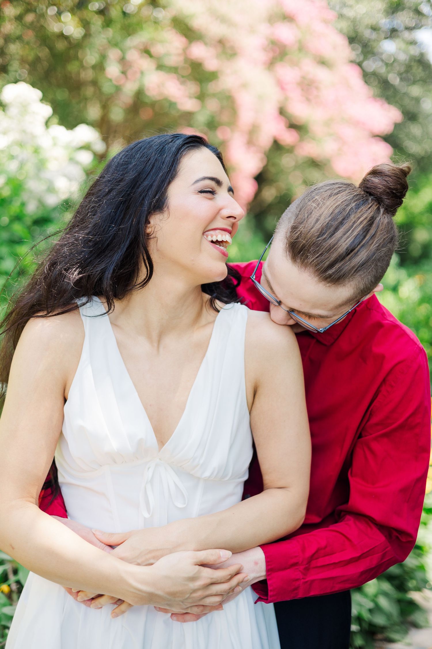 A couple laughs and embraces while posing for romantic photos in a flowering garden.
