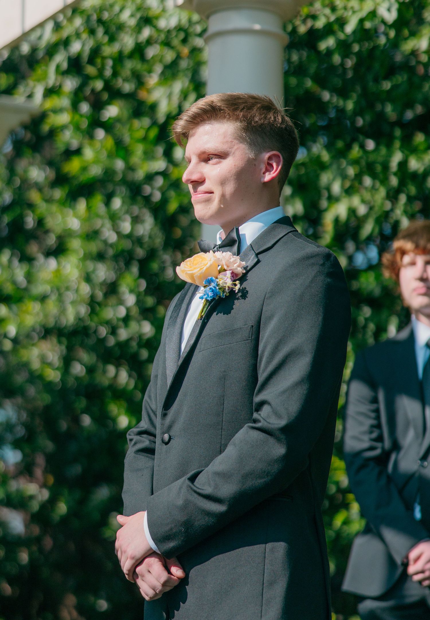 A groomsman in a gray suit with boutonniere stands outdoors with greenery in the background.