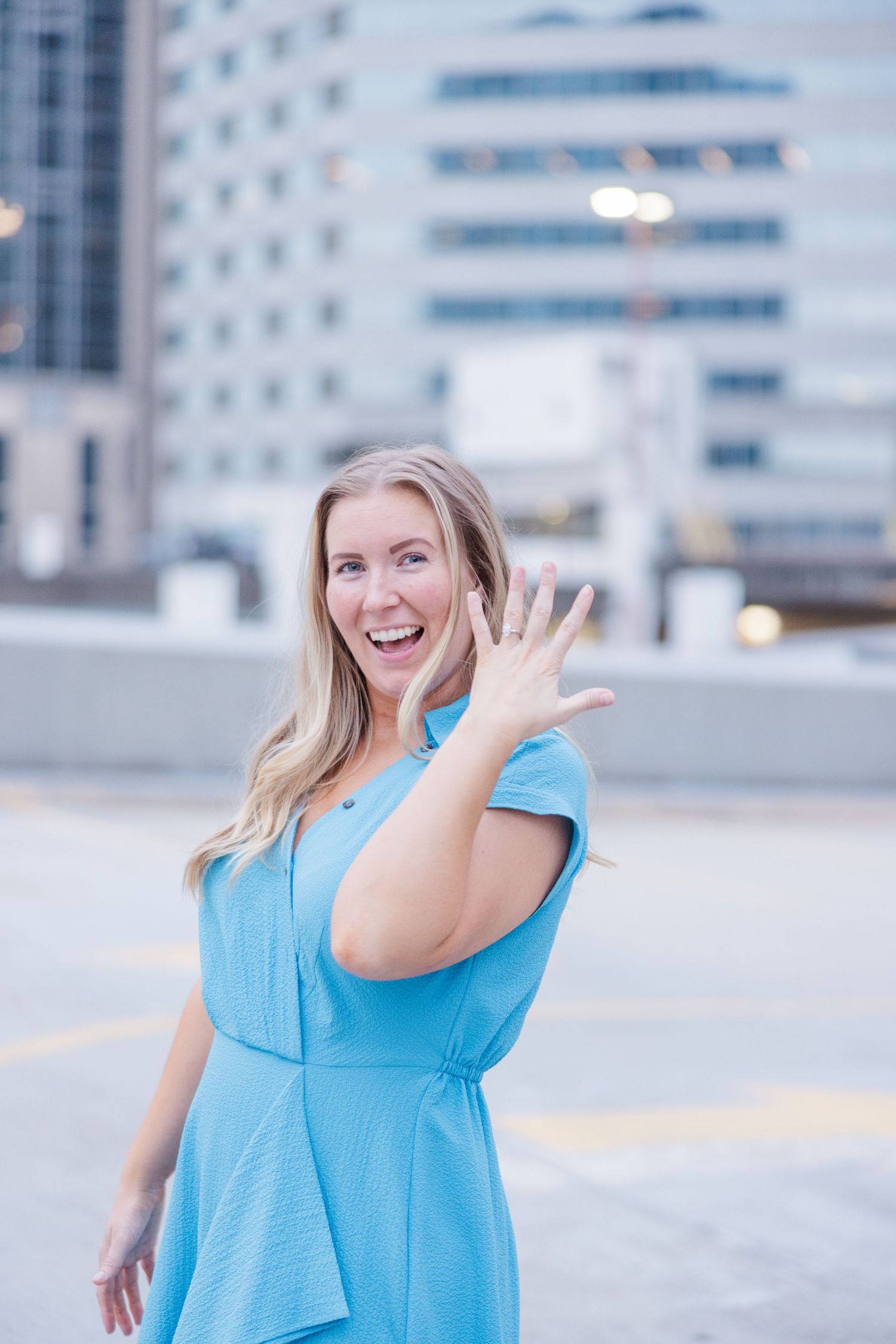 A person in a bright blue top poses expressively against an urban backdrop of modern buildings.