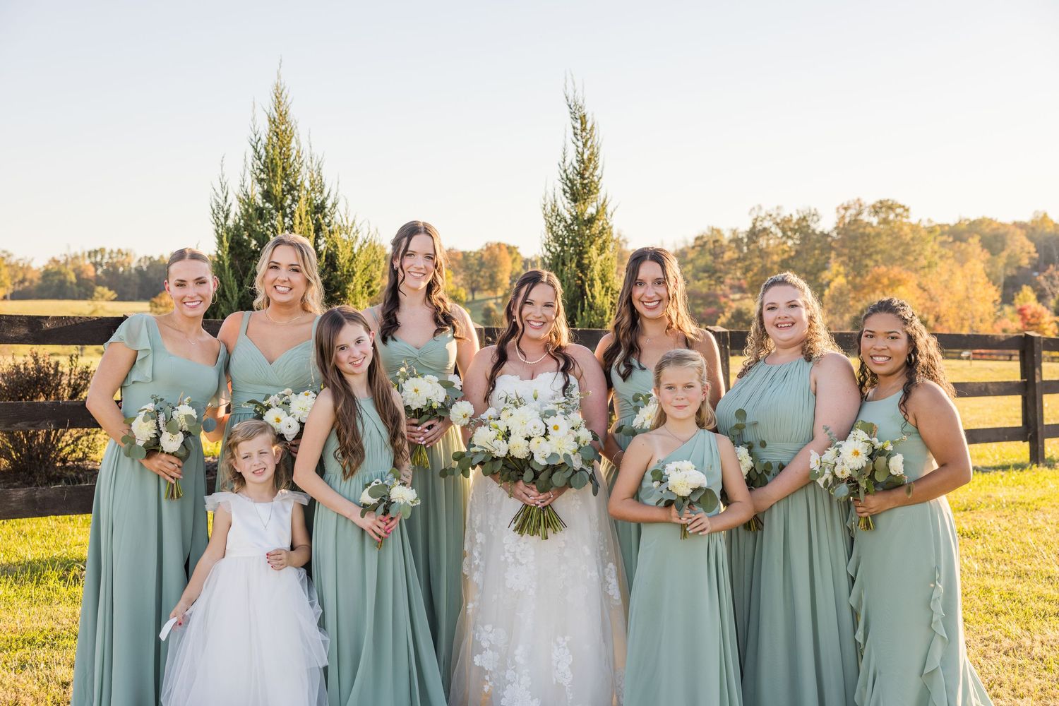 Bridesmaids in sage green dresses holding bouquets pose together outdoors.
