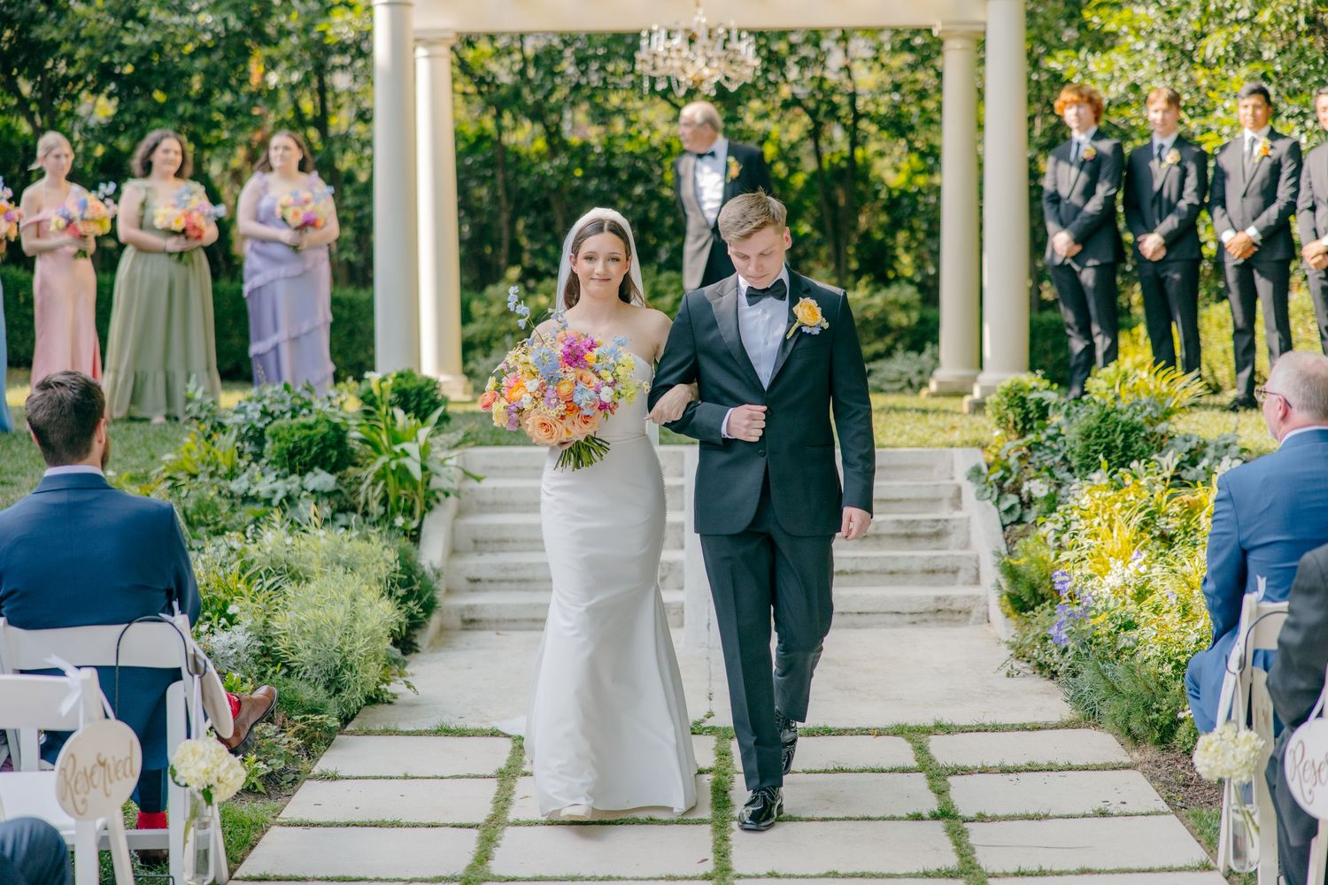 A wedding ceremony processional in a garden setting with bridesmaids in sage dresses and white floral decorations.