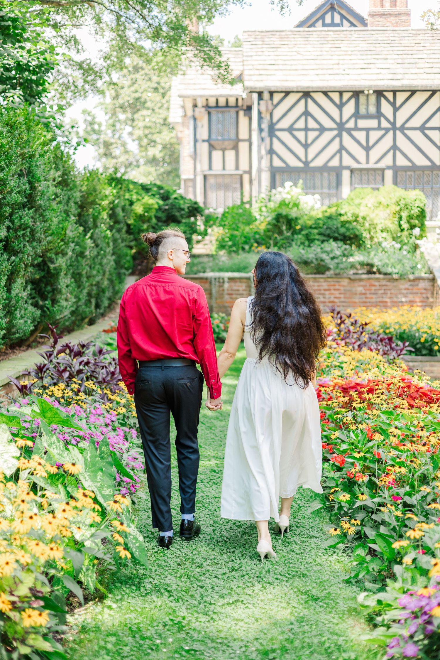 A couple walks hand in hand through a vibrant garden path beside colorful flower beds.