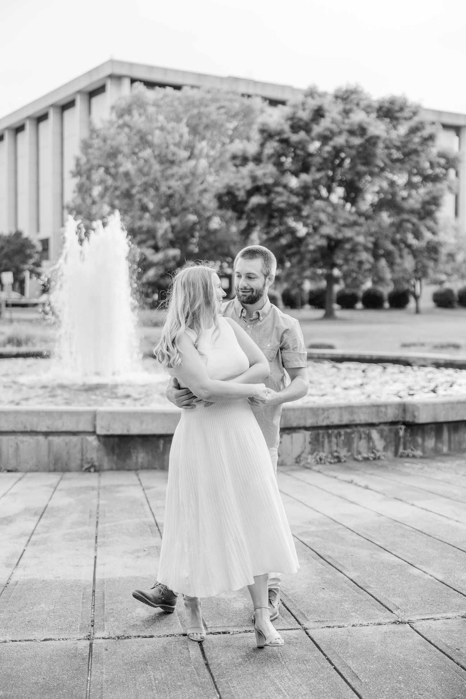 A couple shares an intimate dance moment near a fountain with a building and trees in the background.