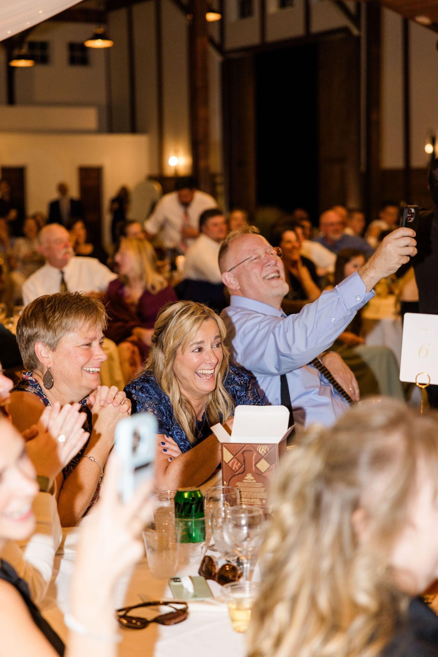 Wedding reception guests laughing and celebrating at tables in an indoor venue.