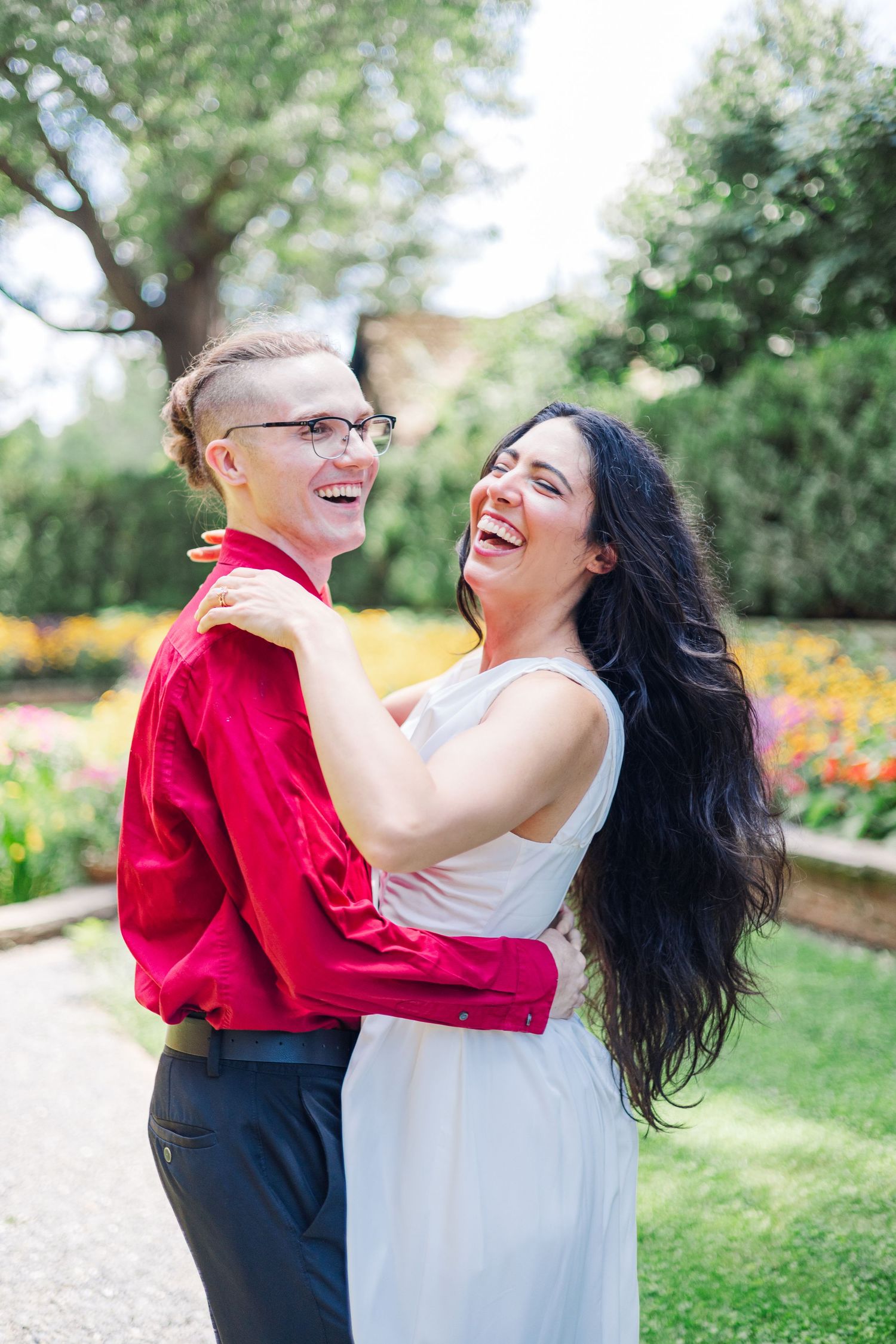 A joyful couple embraces and laughs together in a garden setting with colorful flowers.