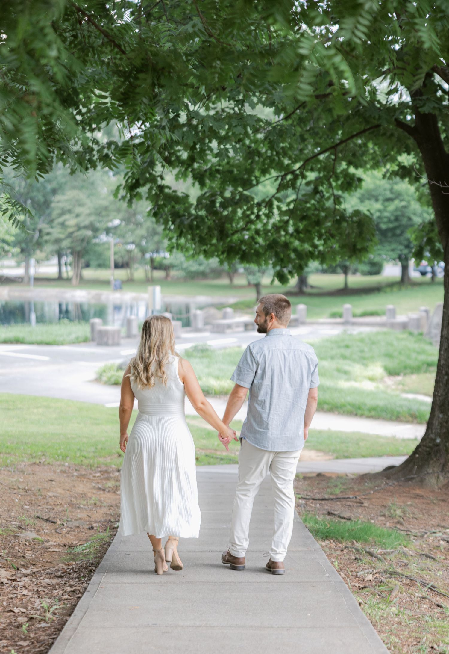 A couple in white outfits walks hand in hand down a peaceful tree-lined path on a summer day.