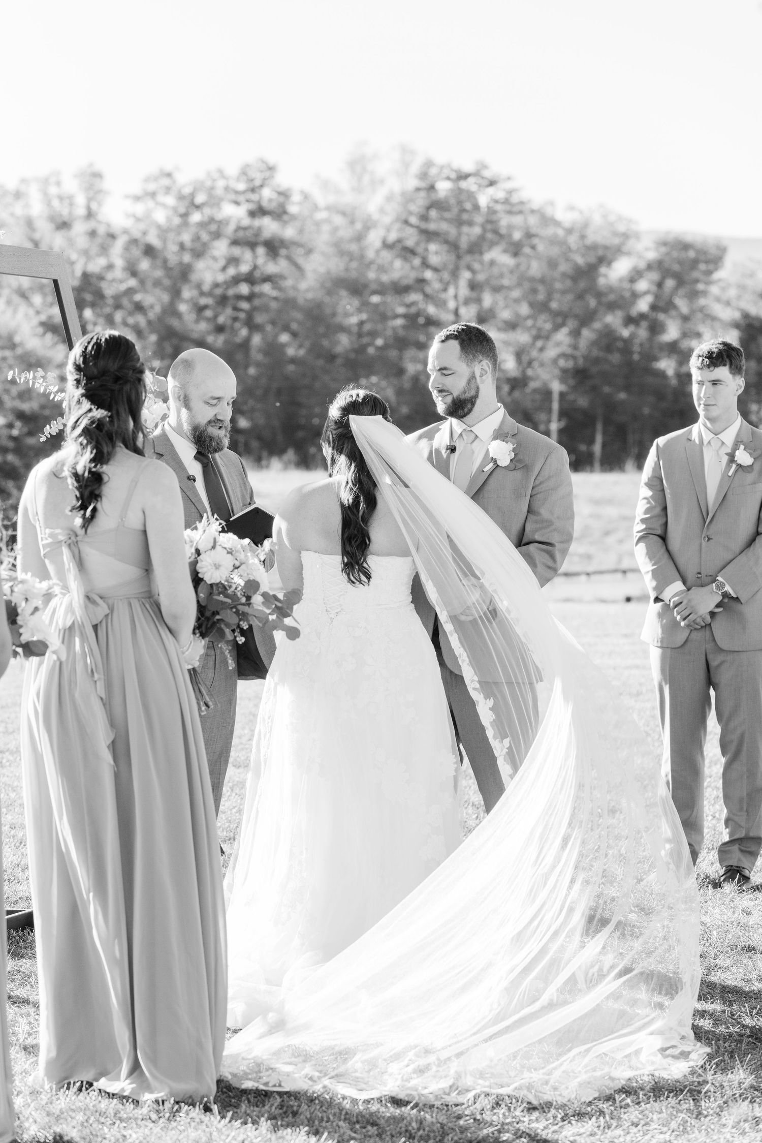 Wedding party in gray attire poses together in a grassy field during an outdoor ceremony.