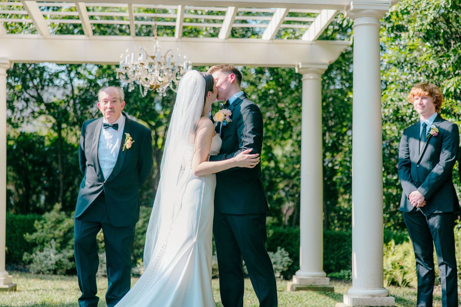 Wedding ceremony under white pergola with crystal chandelier and outdoor garden setting.