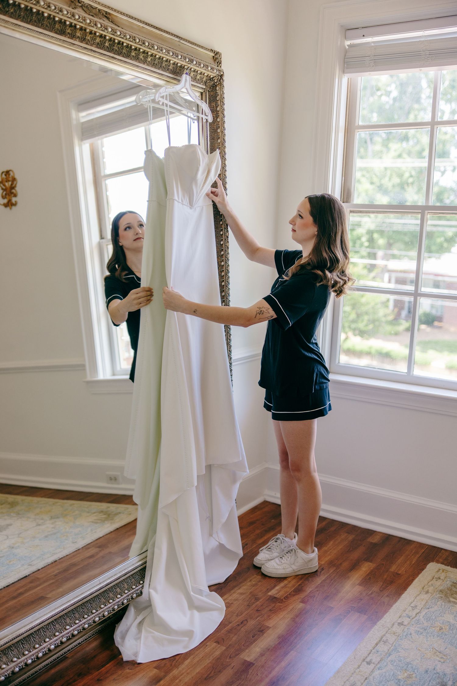 A white wedding dress being held up in front of an ornate mirror in a bright room with hardwood floors.
