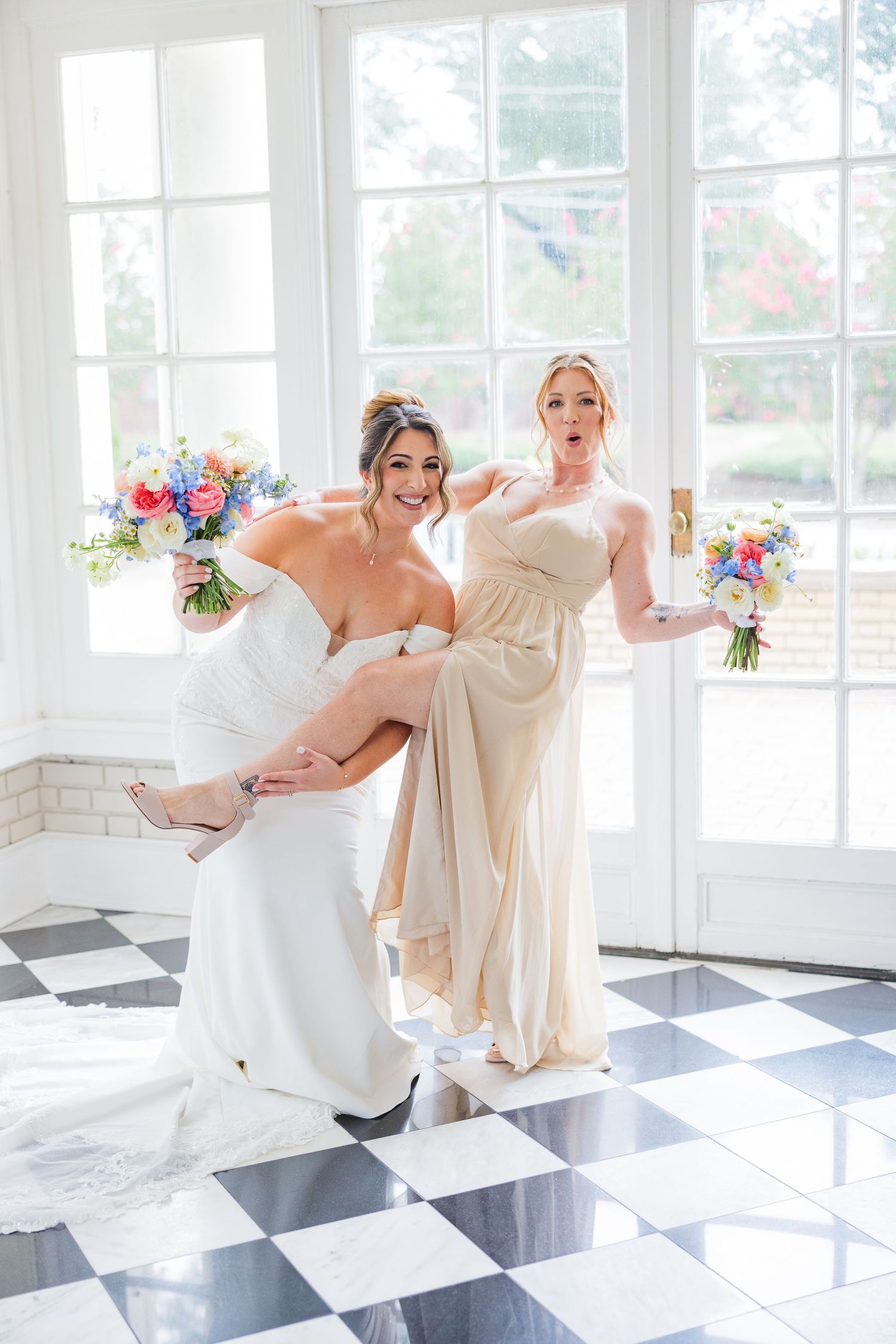 A group in formal attire dances and poses playfully on a black and white checkered floor in a bright sunlit room.