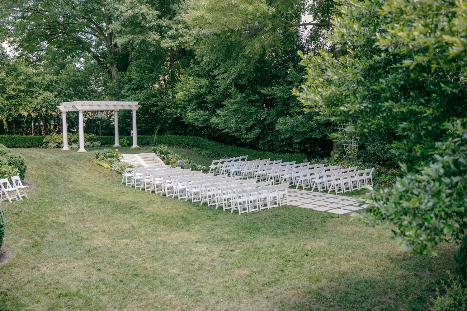 Outdoor wedding ceremony setup with white chairs and gazebo surrounded by lush green trees.