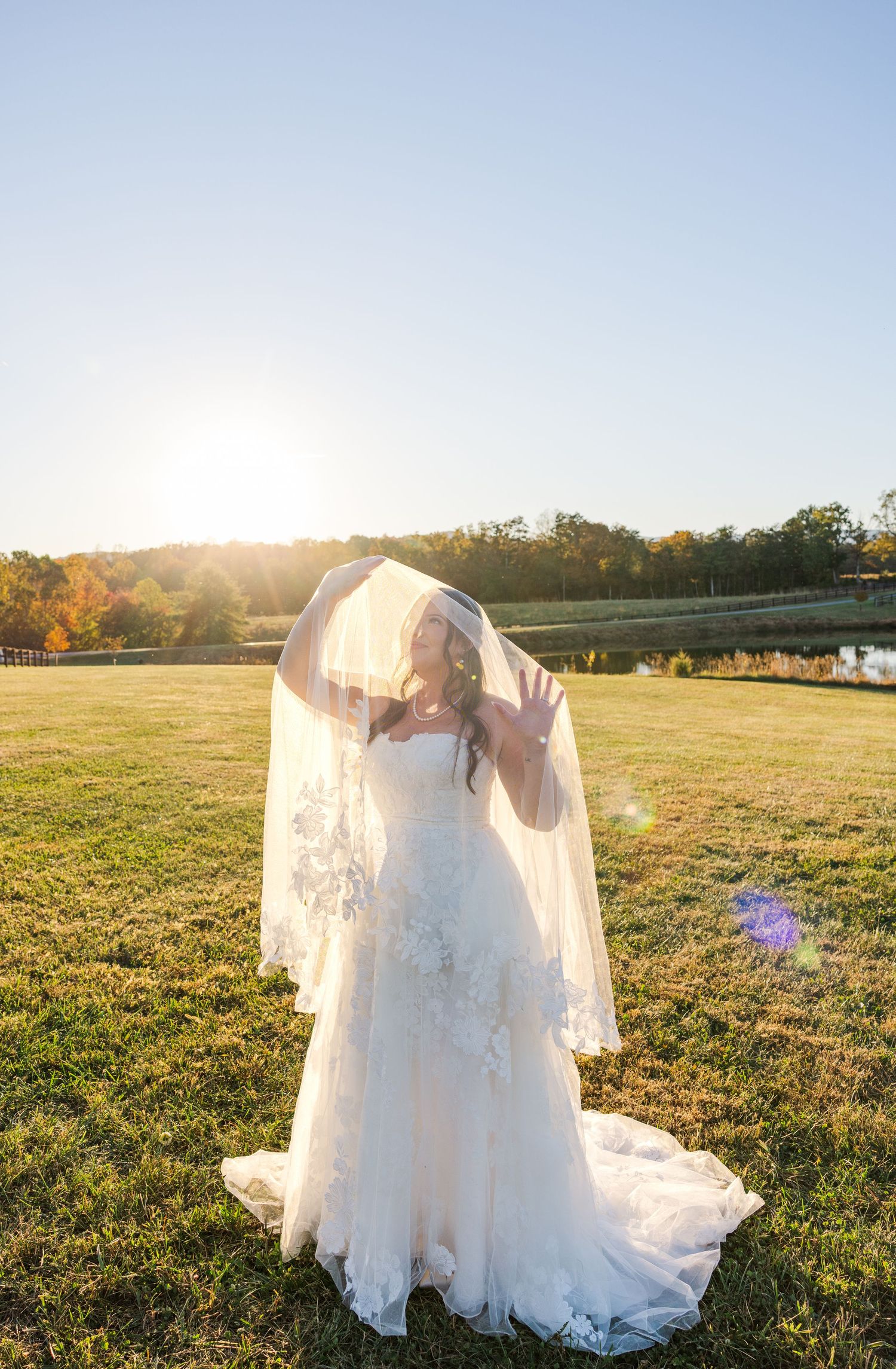 Bride in white lace gown poses in golden sunlit field during sunset.