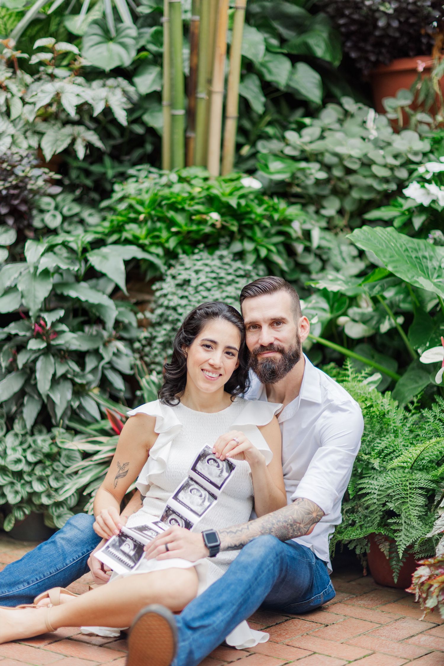A couple relaxes on brick pavement surrounded by lush greenery while holding ultrasound photos.
